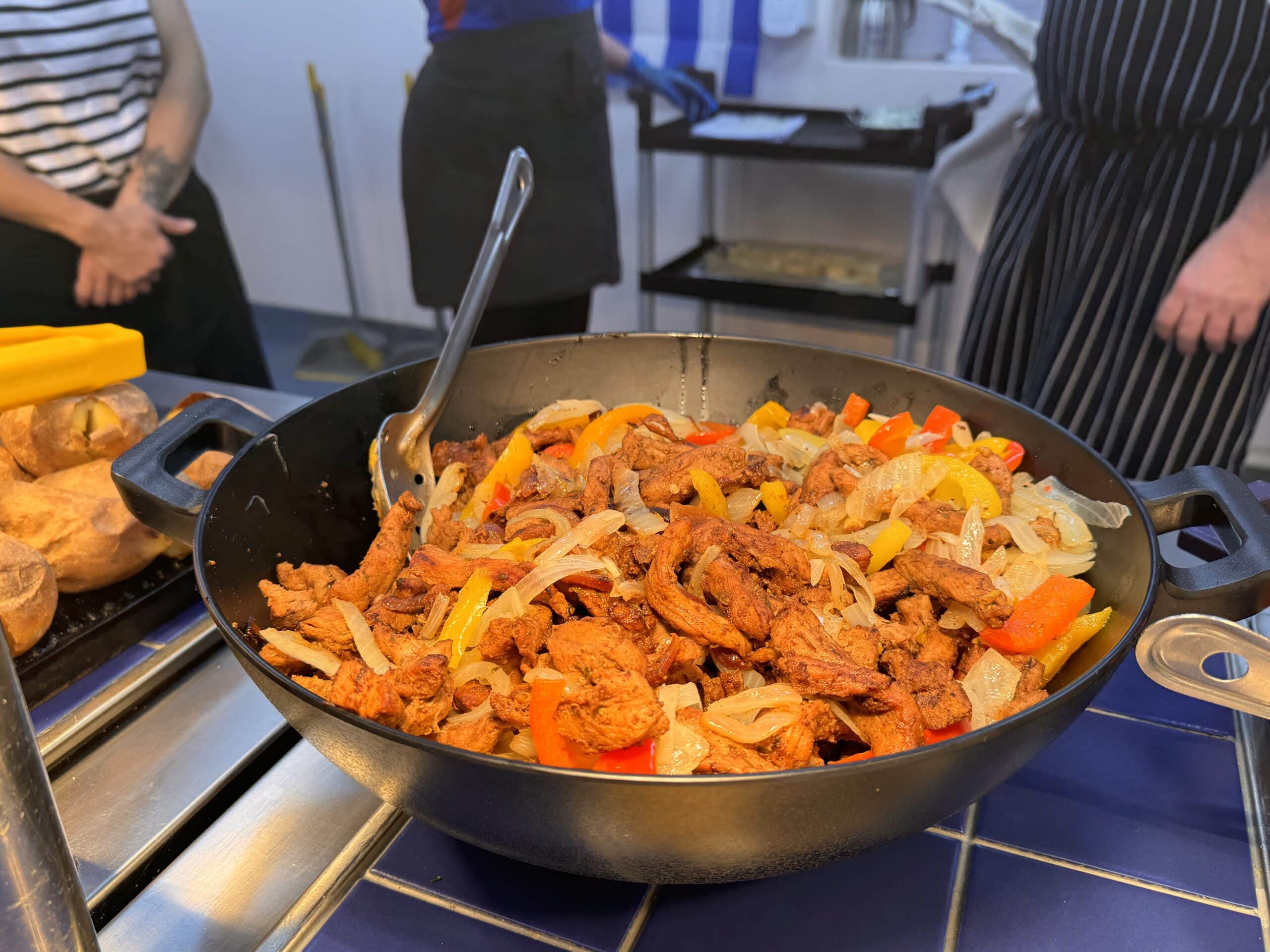 A large black serving pan filled with sizzling strips of spiced chicken, sautéed onions, and red and yellow bell peppers, with a metal serving spoon resting inside. Fresh baked potatoes are visible to the side, with kitchen staff standing in the background.