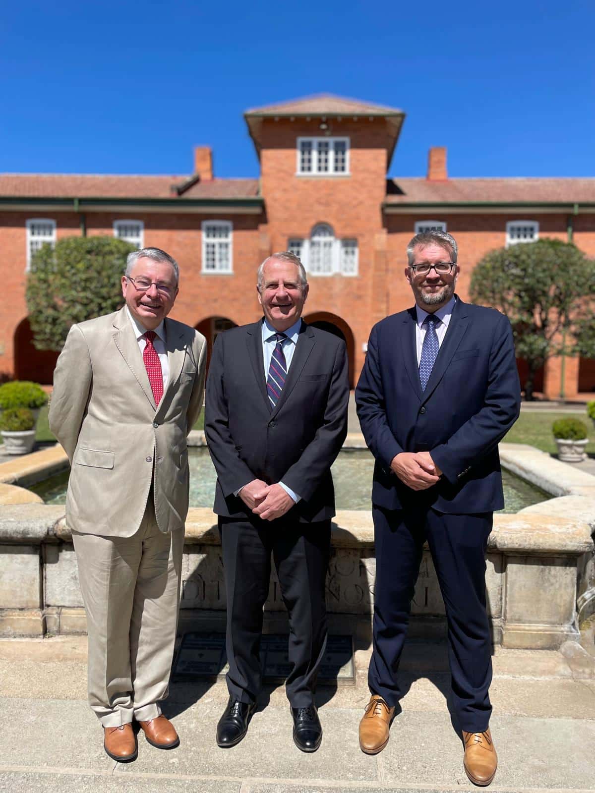 Three men in suits pose in front of a red-brick building with white-trimmed windows and a central tower. They are standing beside a stone fountain under a clear blue sky, suggesting a formal or commemorative occasion.