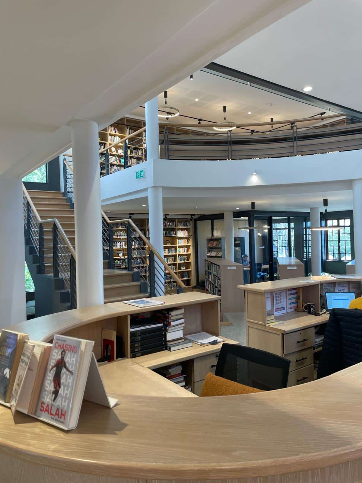 A modern library interior with a curved wooden reception desk, bookshelves on two levels, and staircases leading to an upper floor. Books and displays are neatly arranged throughout the well-lit space.