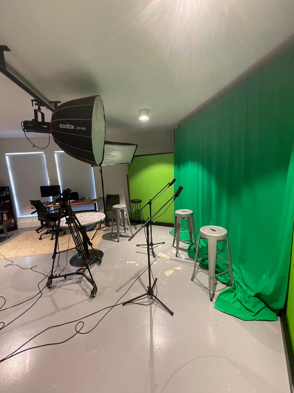 A video production room set up with a green screen backdrop, three tall metal stools, softbox lighting, a camera on a tripod, and multiple microphones on stands. Cables are visible on the light grey floor.