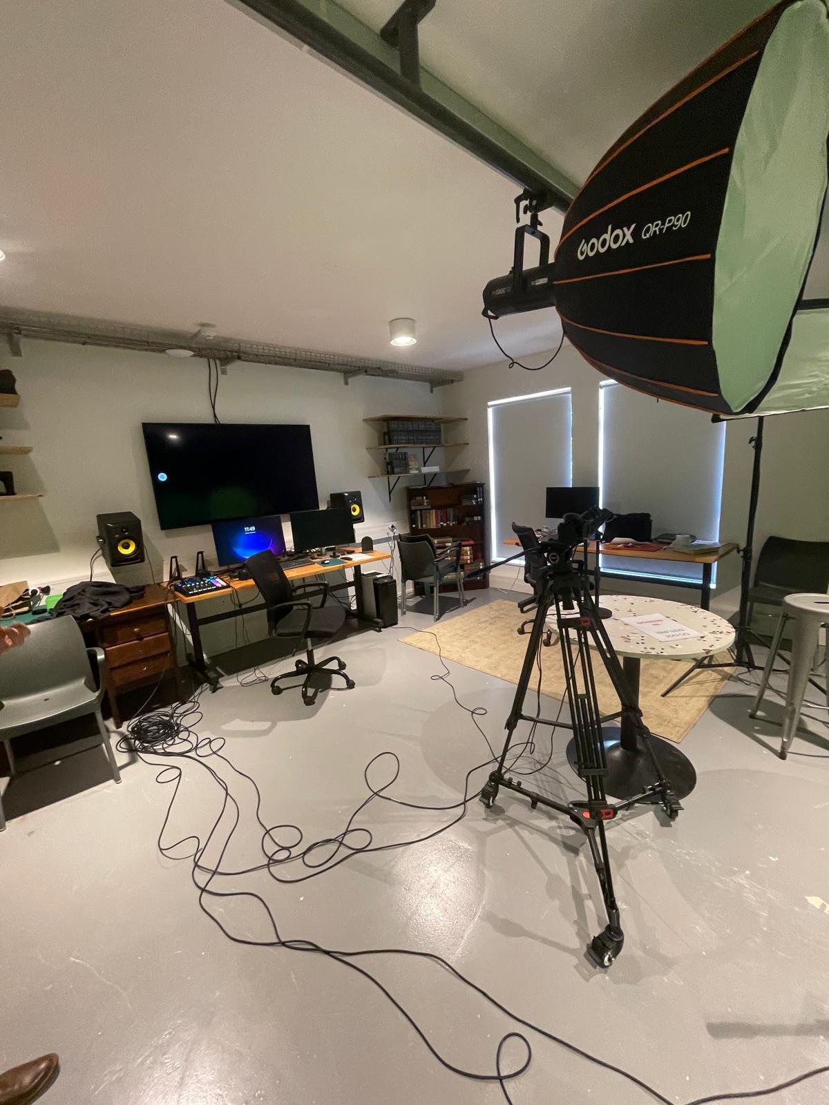 A multimedia studio with video production equipment, including a large camera on a tripod, softbox lighting, computer monitors, speakers, and coiled cables on the floor. The room has a workspace setup with desks and chairs.