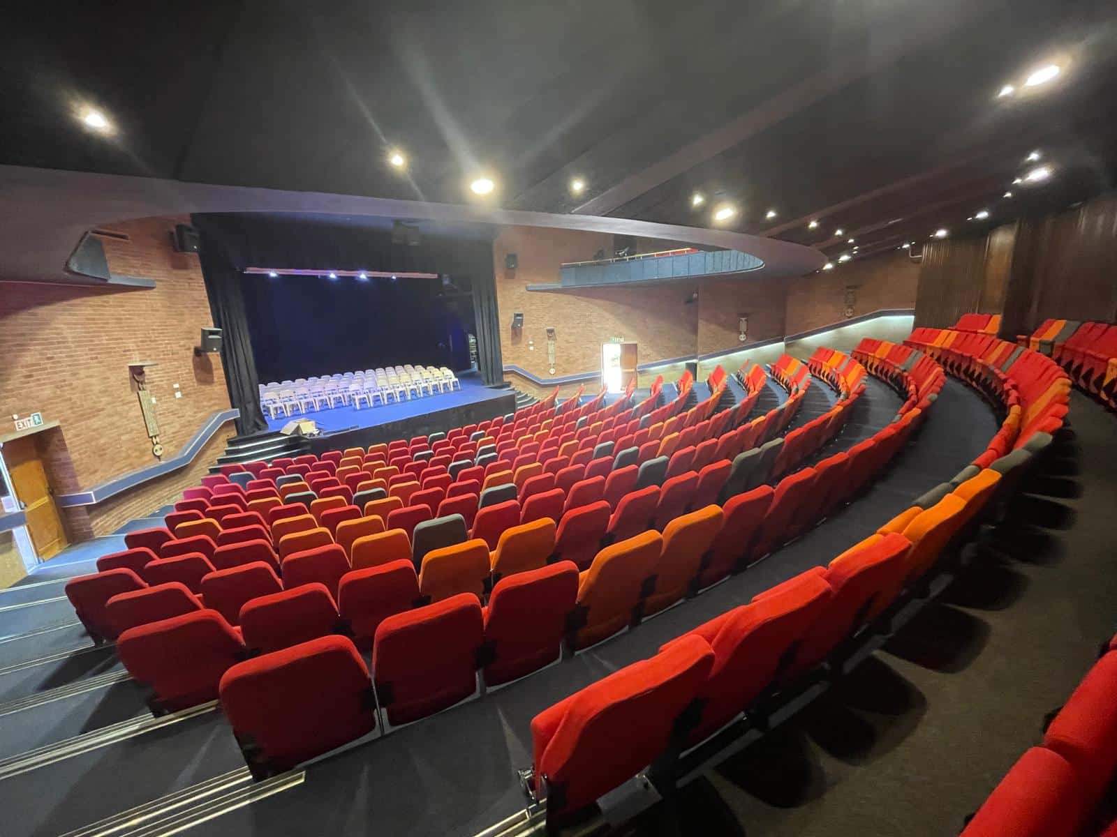 An auditorium with tiered rows of red and orange cushioned seats facing a stage with a row of chairs. The space features curved architecture, brick walls, and overhead lighting.