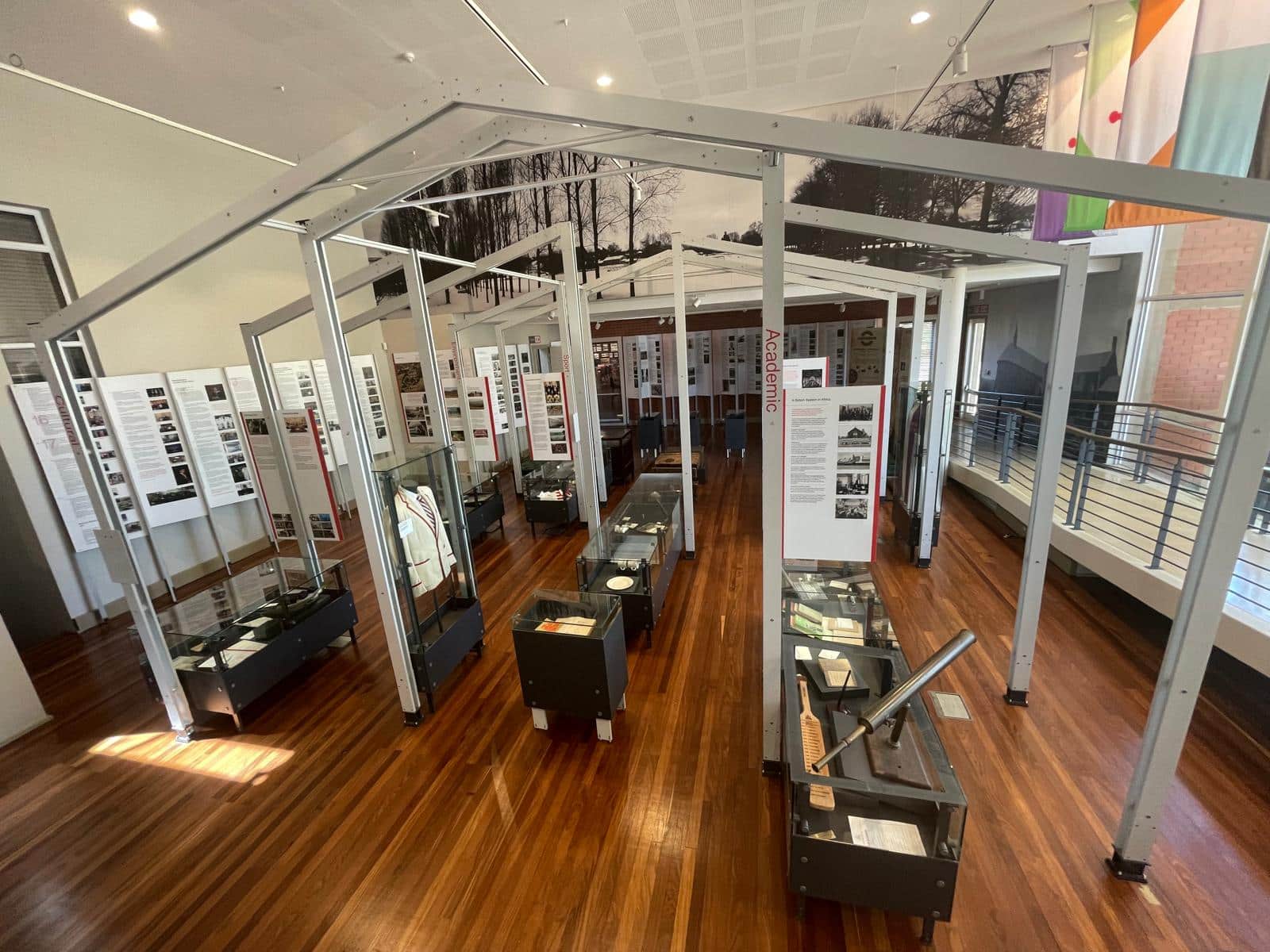 An exhibition space with wooden floors and a modern open-frame structure displaying academic and historical artifacts in glass cases. Large panels with text and photographs line the walls, and banners hang from the ceiling. Natural light enters through windows on the right.