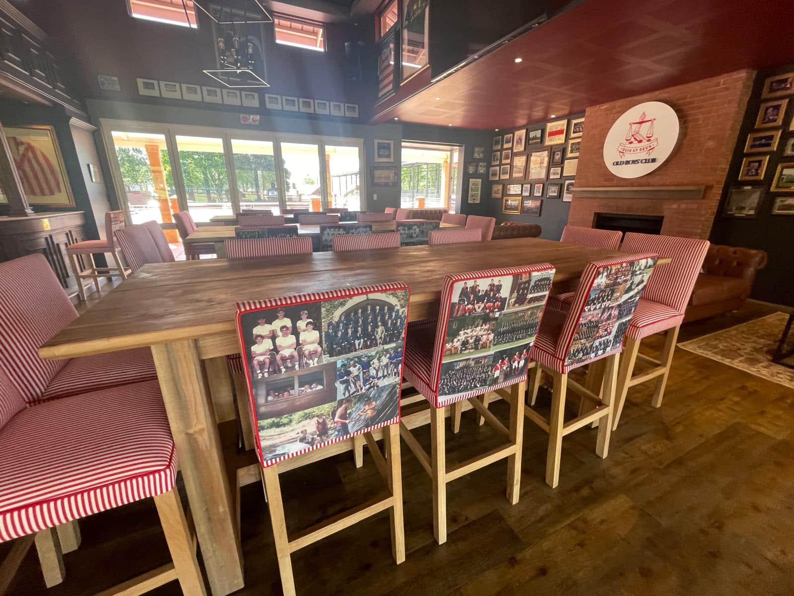 A club-style room with wooden furniture and red-striped chairs, each backrest featuring printed group photos. The room has a cozy, heritage ambiance with framed memorabilia on the walls and a round logo above a brick fireplace reading 
