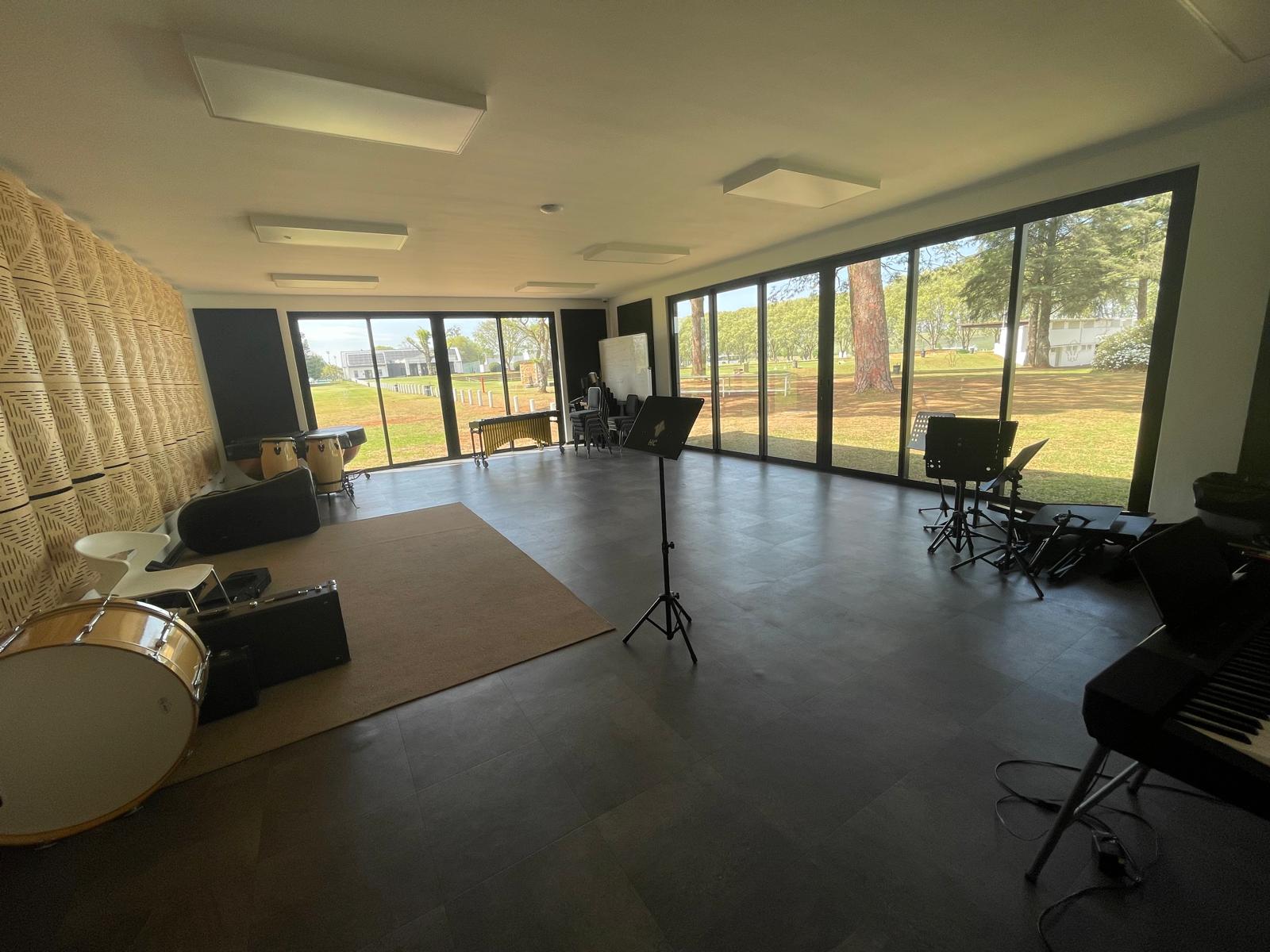 A spacious music room with large floor-to-ceiling windows overlooking a green outdoor area. The room contains percussion instruments, music stands, a keyboard, and an acoustic panel wall on the left for soundproofing.