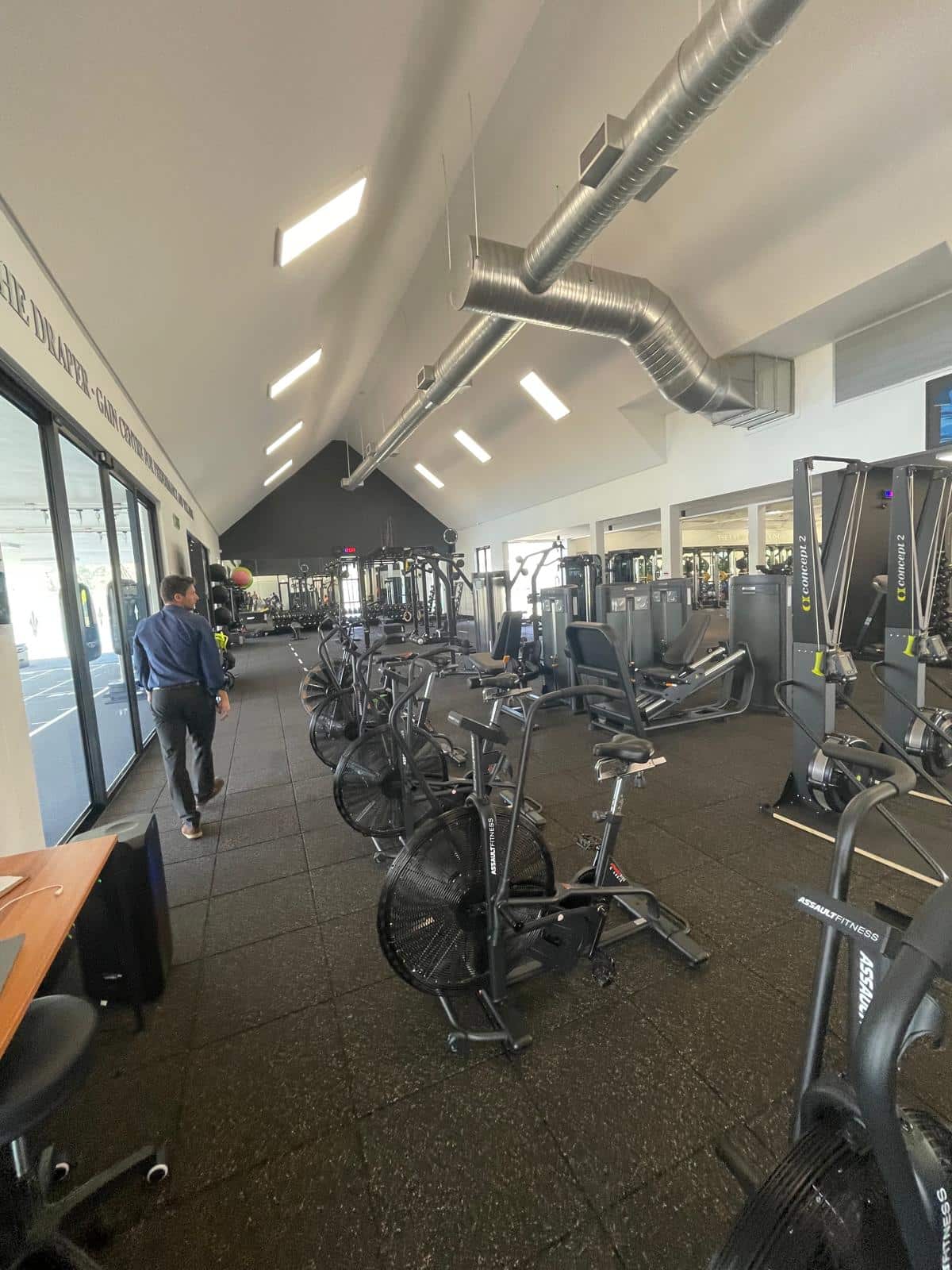 A modern gym with high ceilings, black flooring, and rows of cardio and strength-training equipment, including AssaultFitness bikes and rowing machines. A man walks along the left side near large windows that provide natural light.