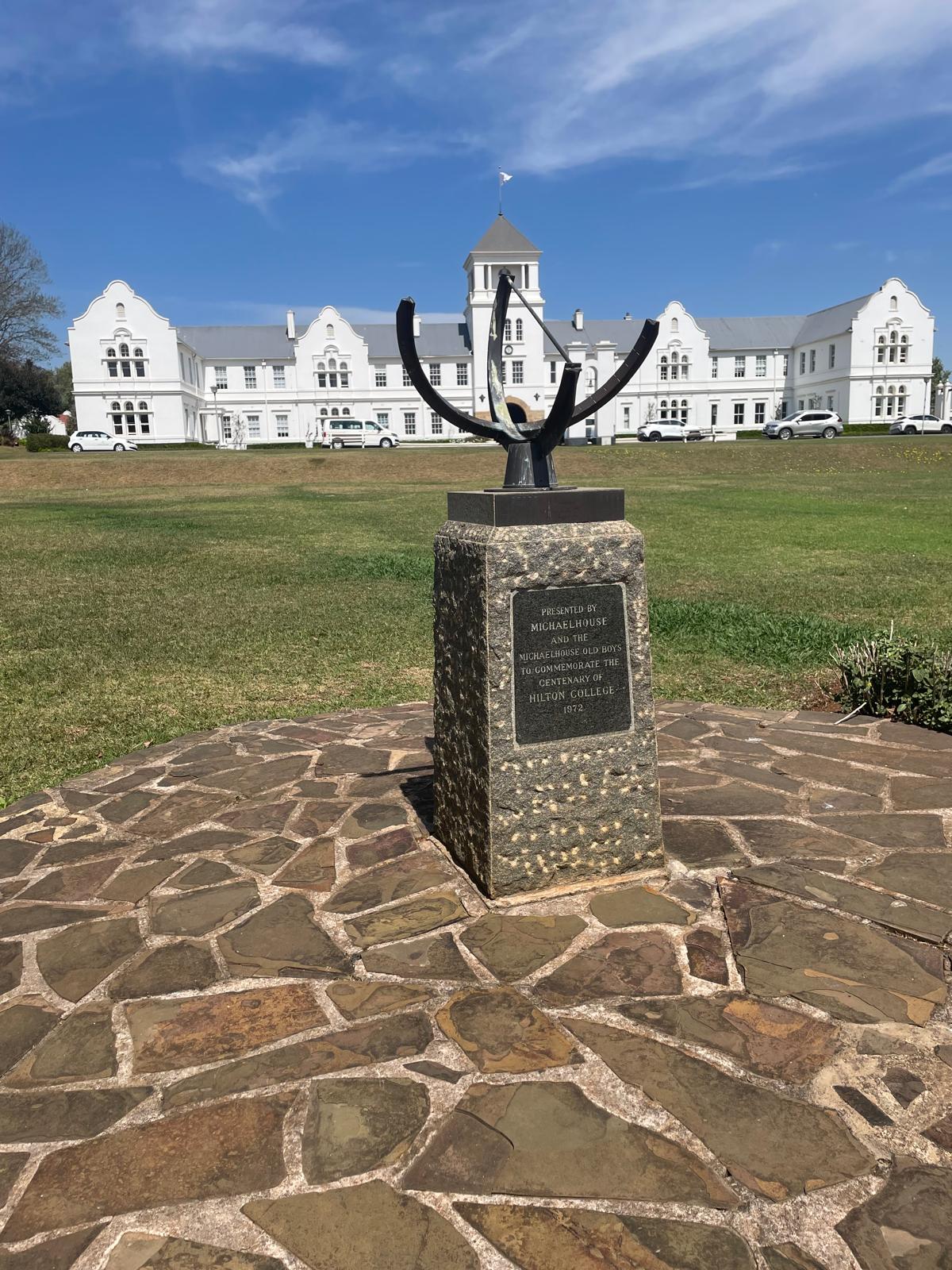 A sundial monument on a stone-paved circular platform, with a plaque commemorating the centenary of Hilton College. In the background stands a large white colonial-style building with a central tower and gabled wings, set against a bright blue sky.