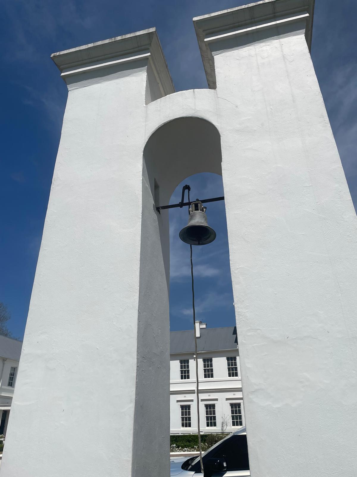 A tall white bell tower with a central arch and a hanging bell, set against a blue sky. A colonial-style white building with large windows and a grey roof is visible in the background, along with a parked car partially in view.