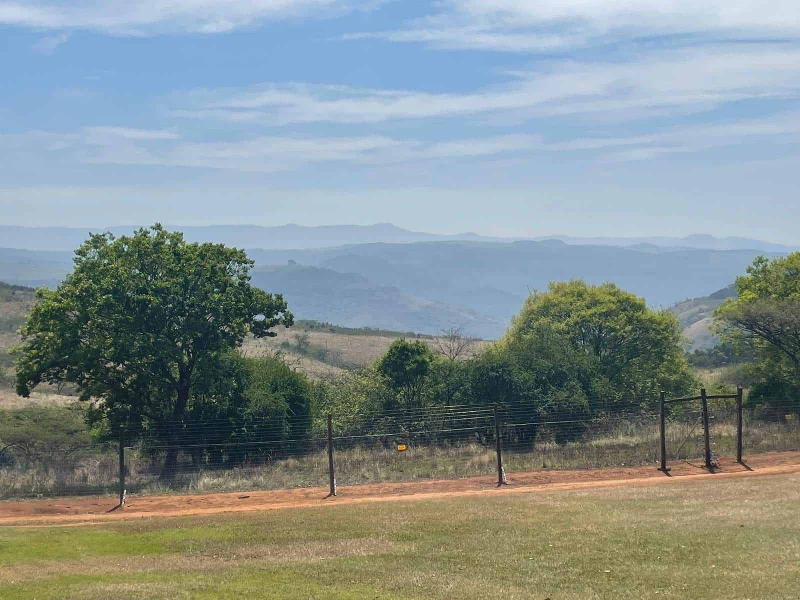 A scenic landscape view of rolling hills and layered mountain ranges in the distance, with scattered green trees and a wire fence in the foreground under a partly cloudy blue sky.