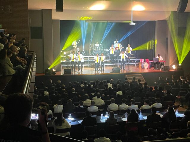 A lively stage performance featuring a group of singers in white shirts and yellow scarves, backed by a band with drums, guitars, and keyboard. Colorful stage lighting with yellow beams highlights the performers, while a large audience, mostly in formal attire, watches from tiered seating in a darkened auditorium.