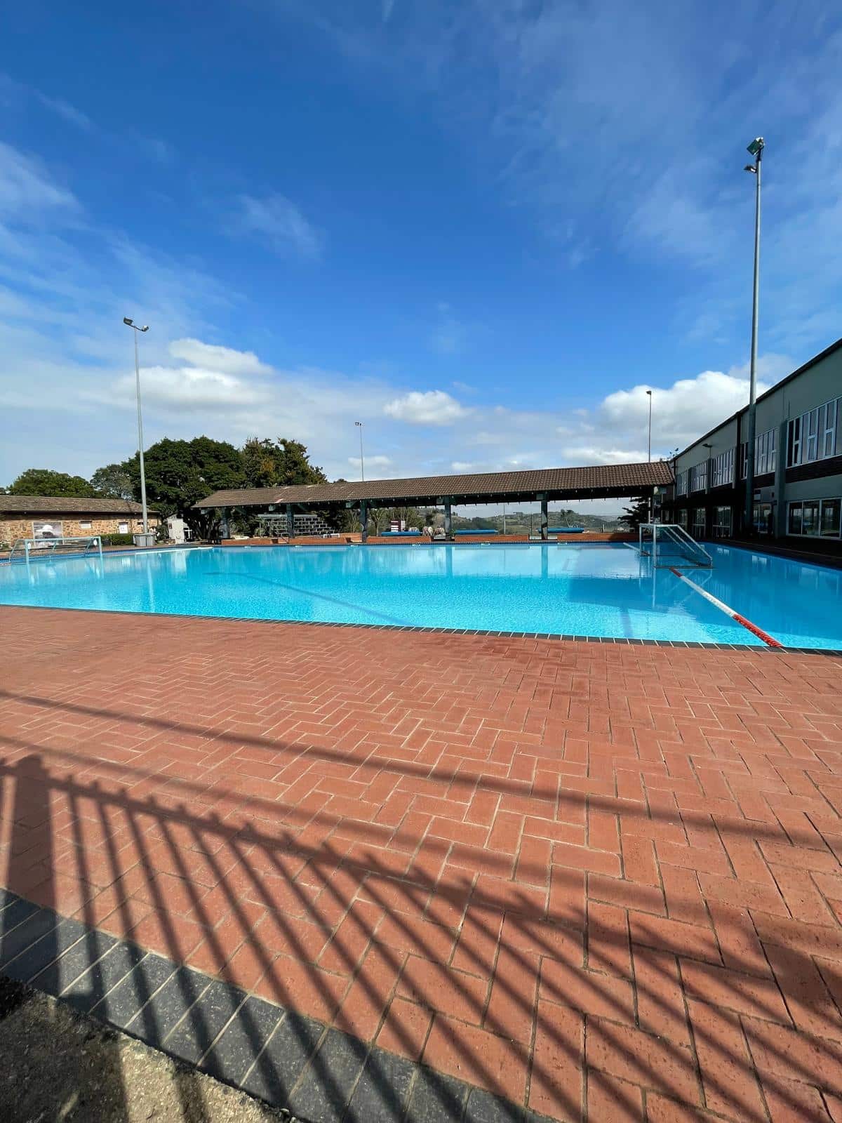 An outdoor swimming pool with clear blue water surrounded by red brick paving. The area is enclosed by fencing, with nearby buildings and a covered seating or viewing area visible in the background under a bright blue sky.