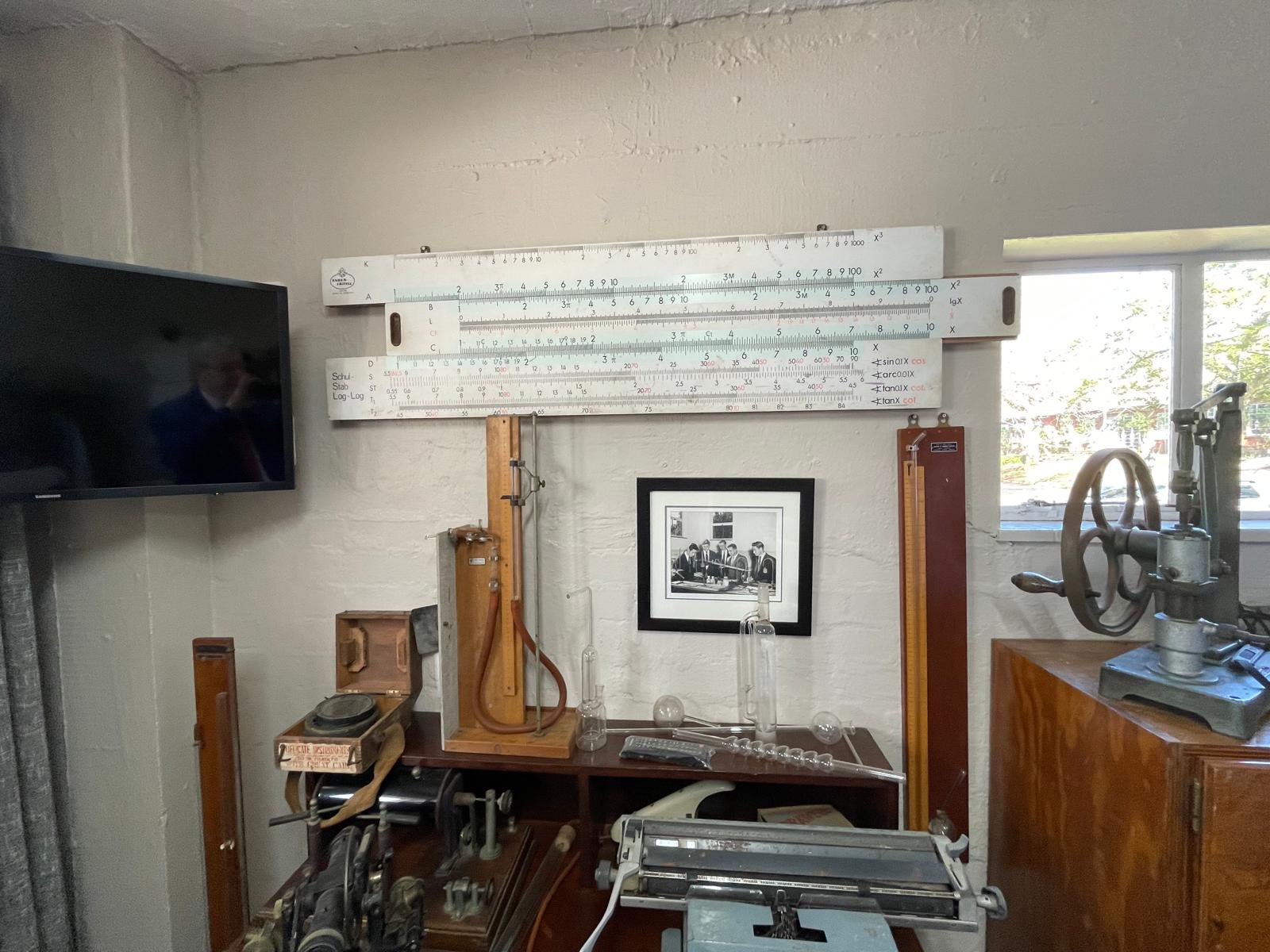 A display of vintage scientific and mechanical instruments in a museum room, featuring a large wall-mounted slide rule, a framed black-and-white photo of men in suits, and various lab equipment including glassware, a typewriter, and a mechanical press. A flat-screen TV is mounted on the adjacent wall.