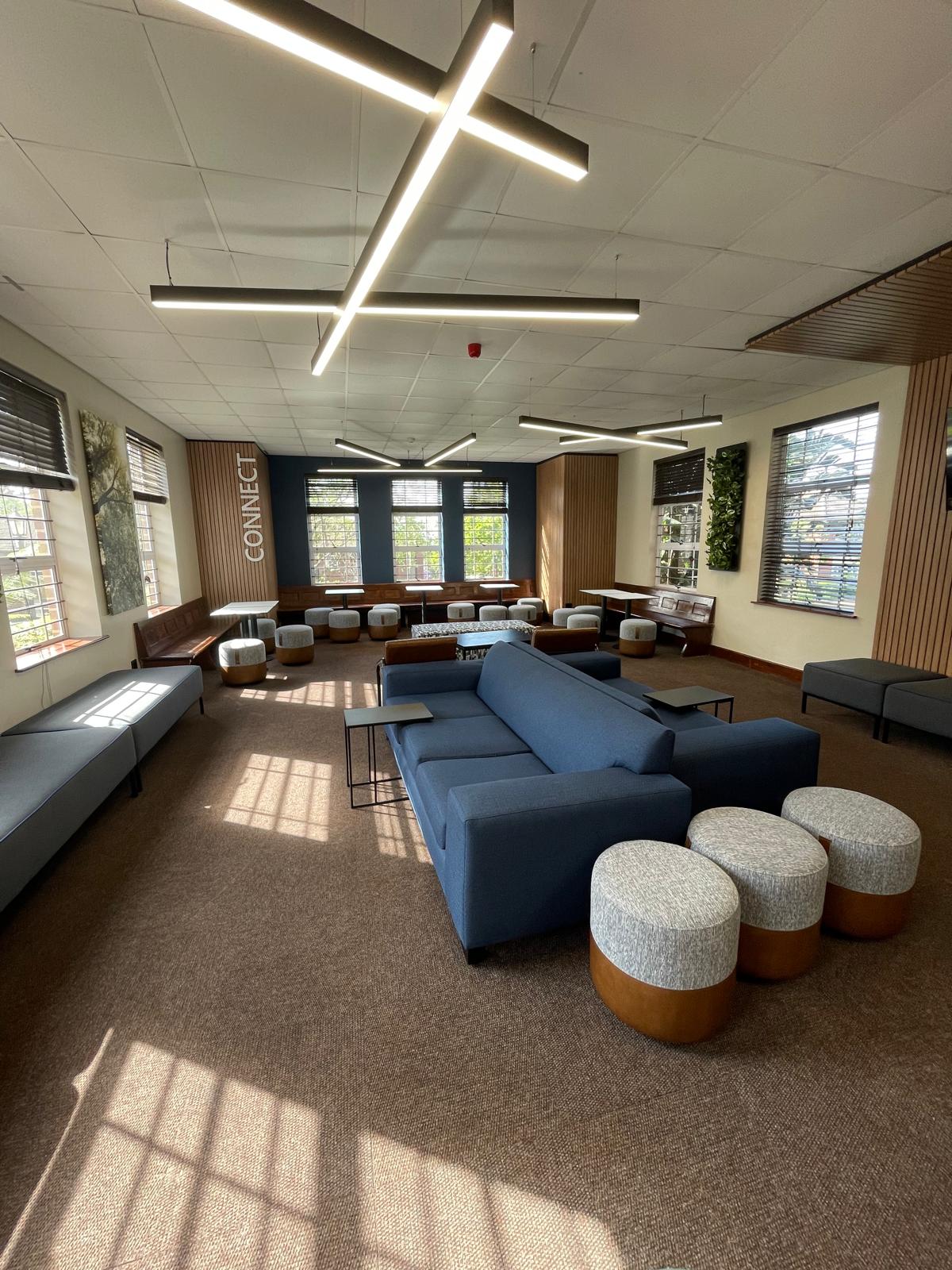 A modern lounge or common room with a mix of seating, including blue sofas, circular ottomans, and benches. Stylish cross-shaped ceiling lights and large windows let in natural light. The word “CONNECT” is displayed on a back wall.