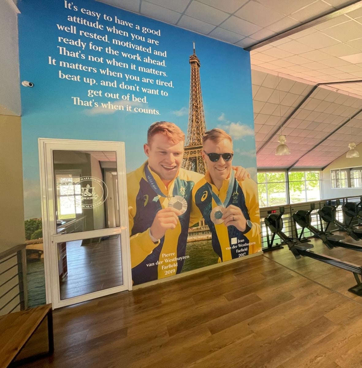 A gym interior with several rowing machines aligned on a wooden floor. A large motivational mural of two medal-winning athletes with the Eiffel Tower in the background includes the names “Pierre van der Westhuyzen” and “Jean van der Westhuyzen,” and a quote about perseverance and attitude.
