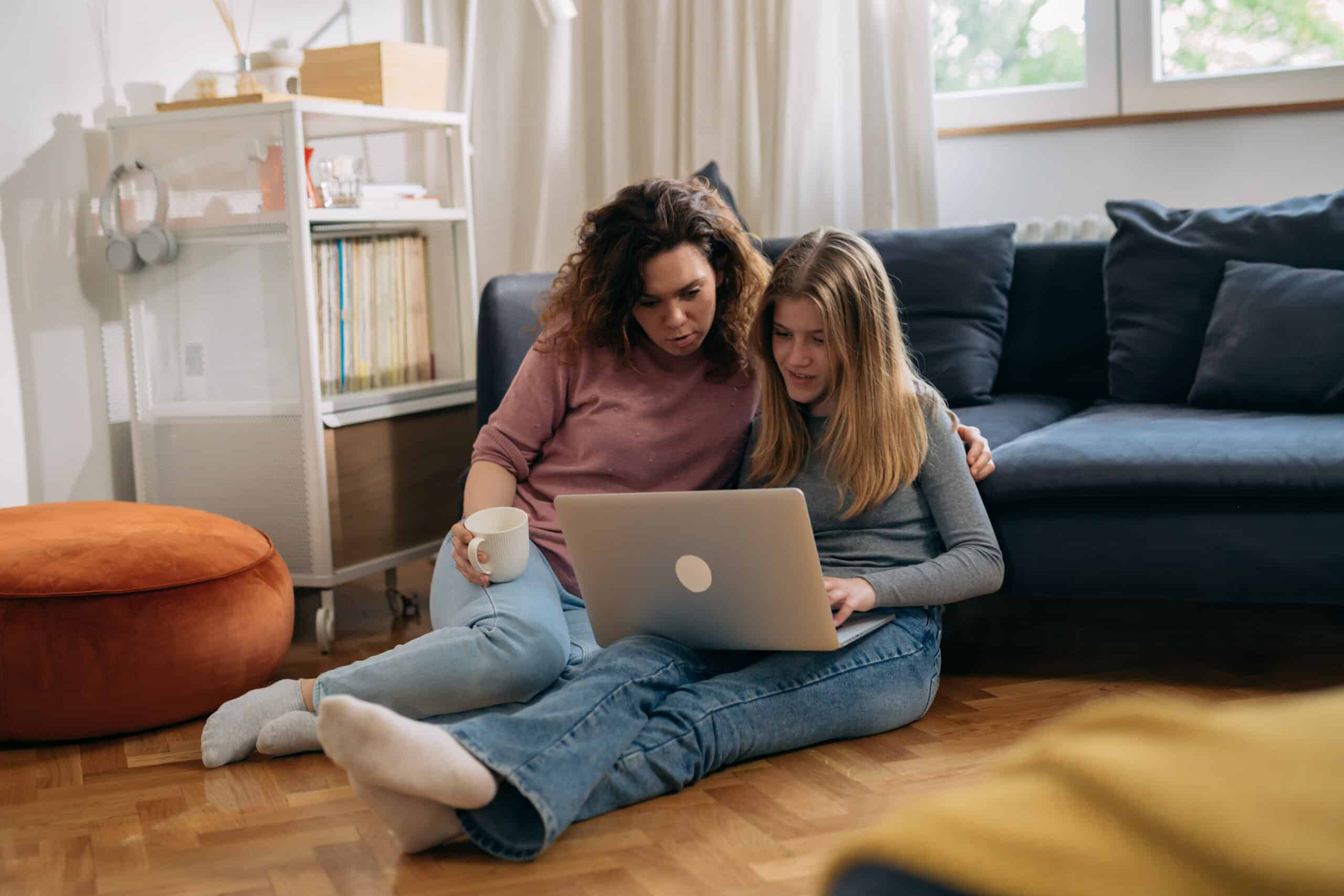 A woman and a teenage girl sitting on the floor in a living room, looking at a laptop together. The woman holds a white mug while the girl types, and they appear engaged in what’s on the screen. A dark sofa, bookshelves, and a large window are visible in the background