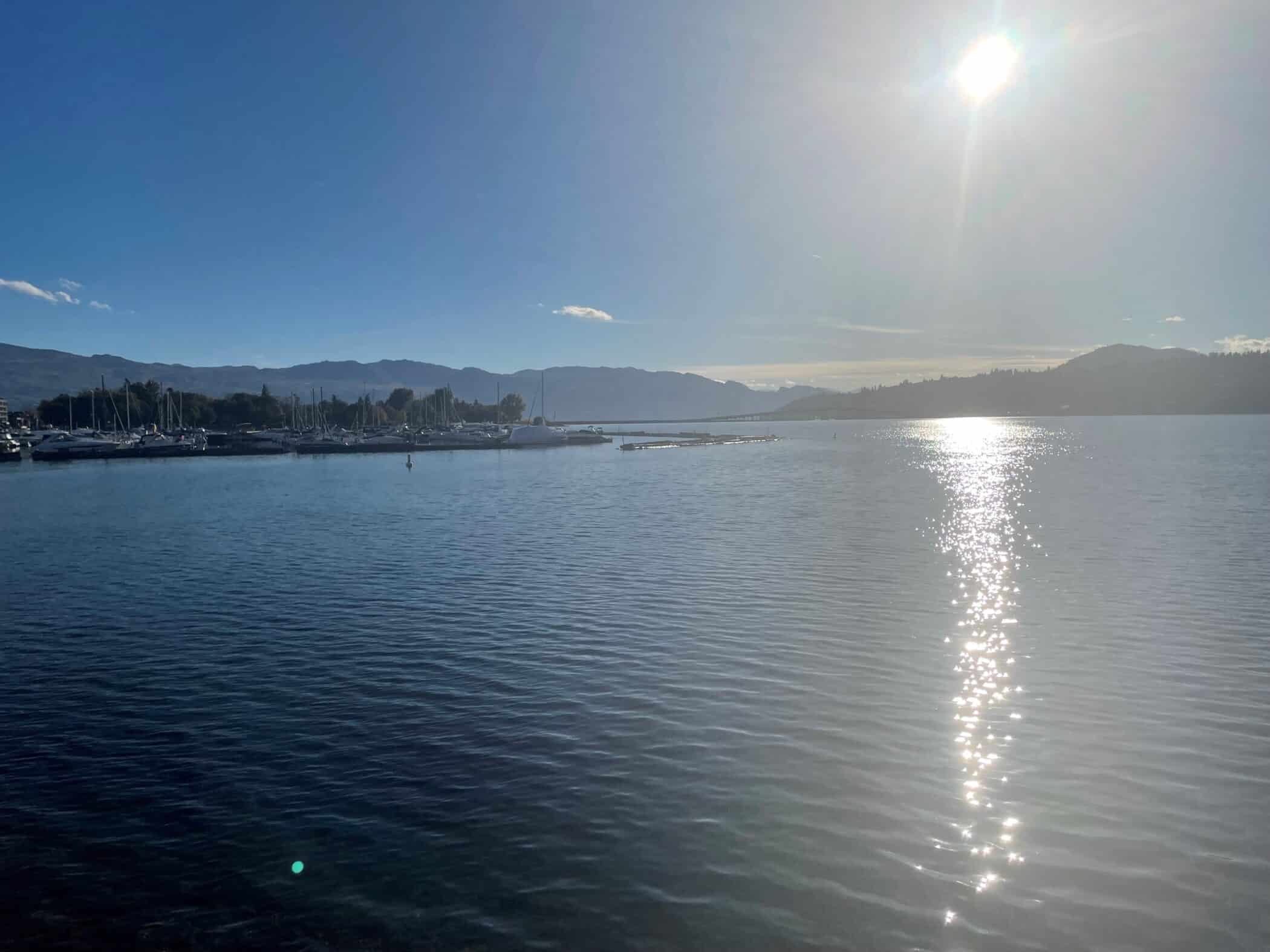 Sunny afternoon view across Lake Okanagan with sailboats docked near the marina and the sun reflecting brightly on the rippling water.