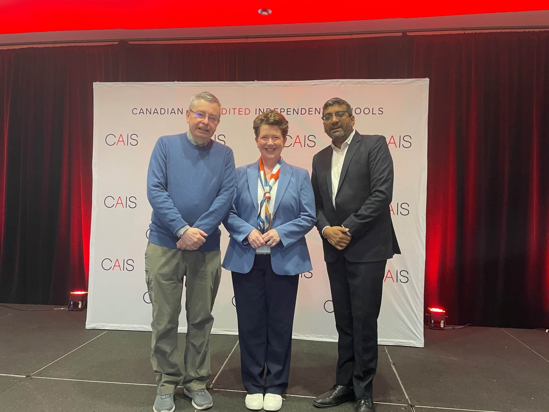 Three people stand on stage in front of a CAIS backdrop smiling for a photo—two men and one woman, all dressed in business or semi-formal attire.