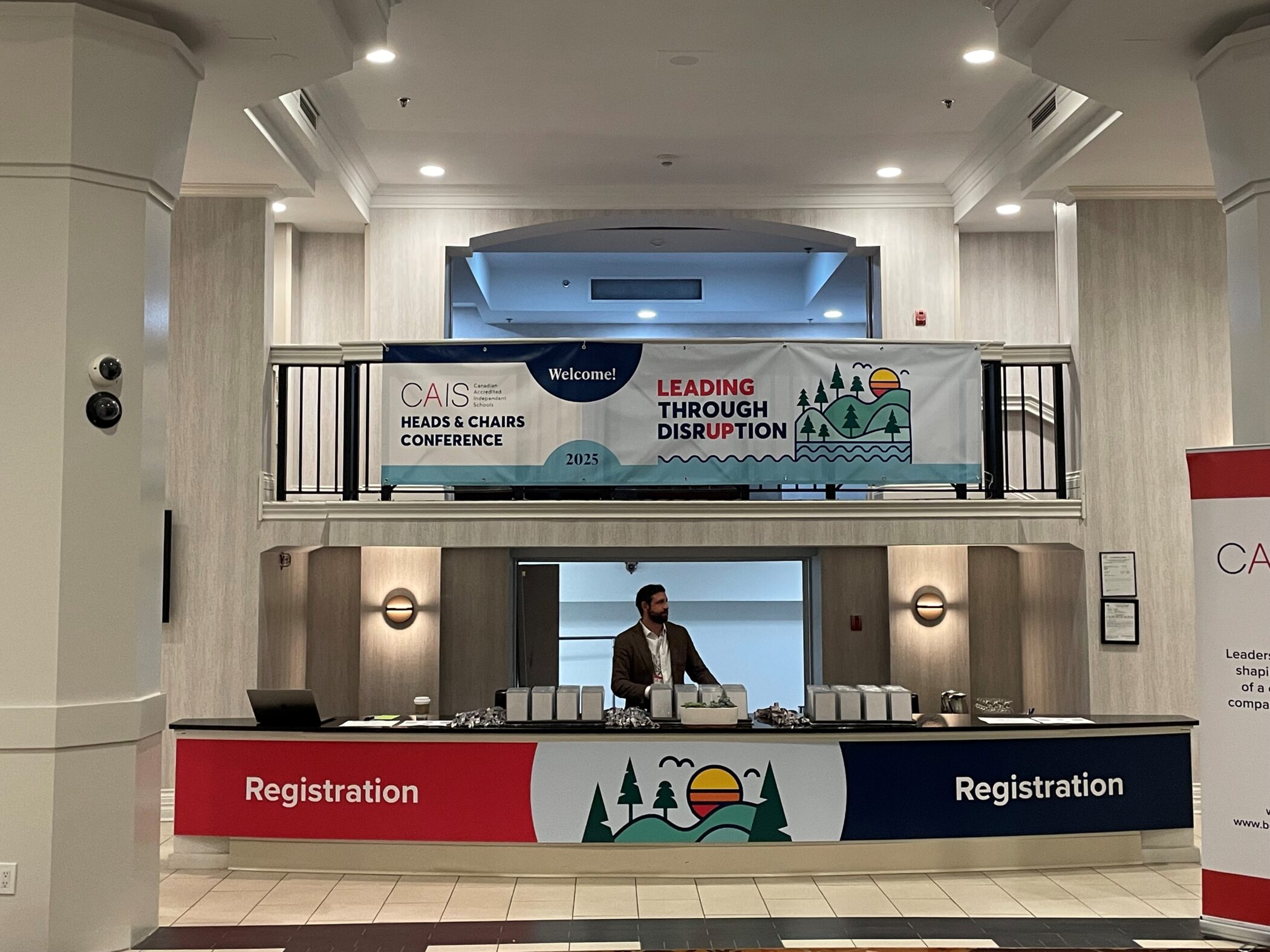 Registration area for the CAIS Heads & Chairs Conference 2025 with a banner reading “Leading Through Disruption.” A man stands behind the registration desk in a hotel lobby.