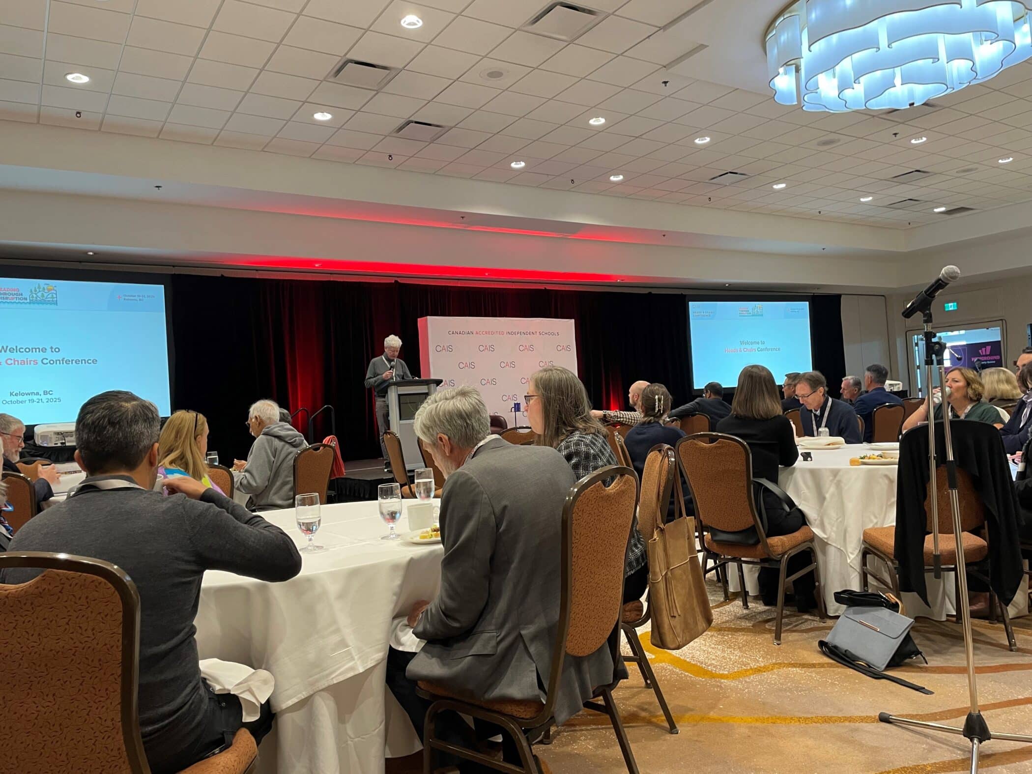 Conference attendees seated at round tables listening to a speaker at the CAIS Heads & Chairs Conference in Kelowna, BC. Two large screens and red stage lighting are visible.
