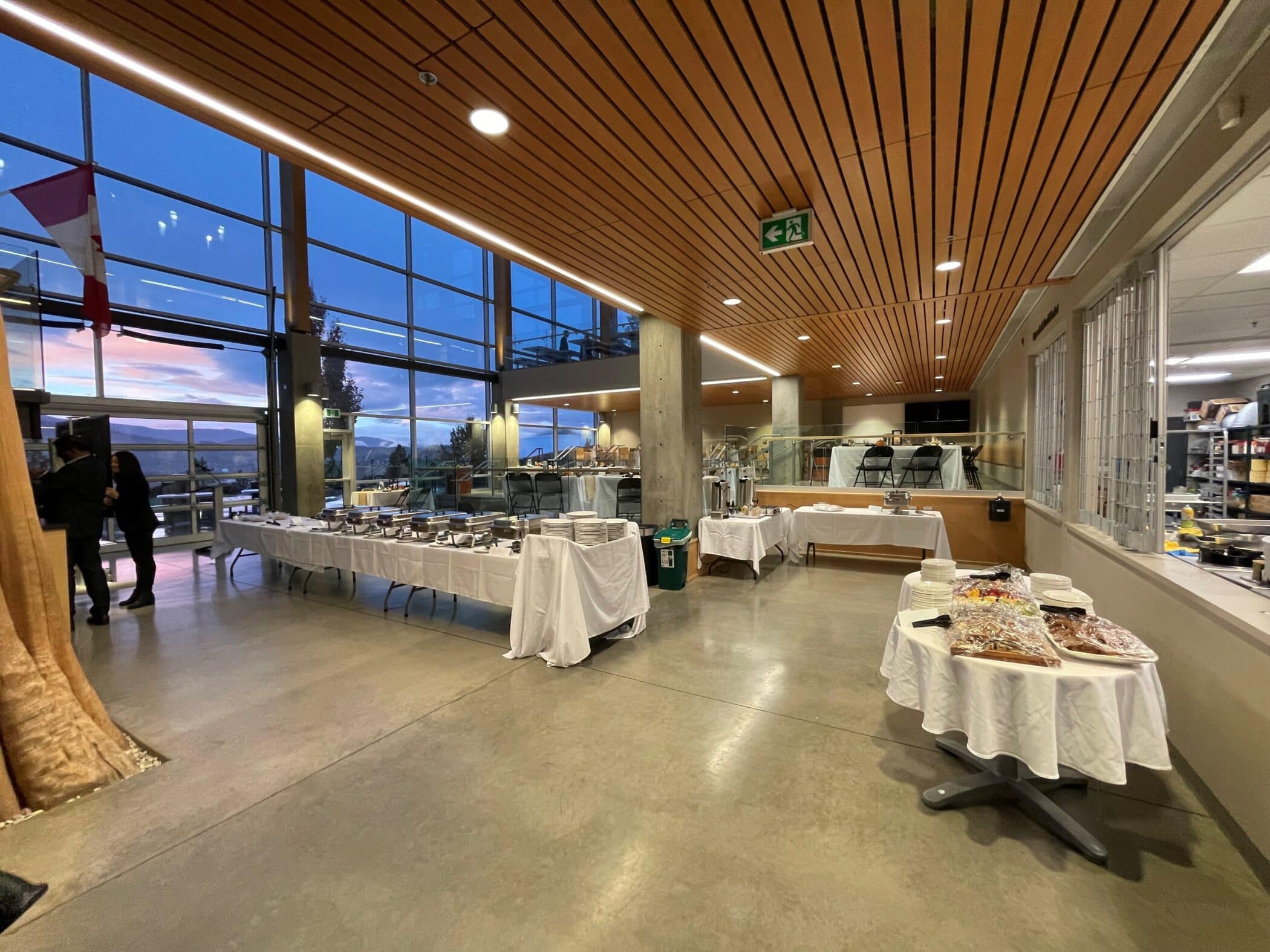 A buffet dinner setup in a modern hall with large windows showing an evening sky. Tables are laid with plates, cutlery, and food displays.