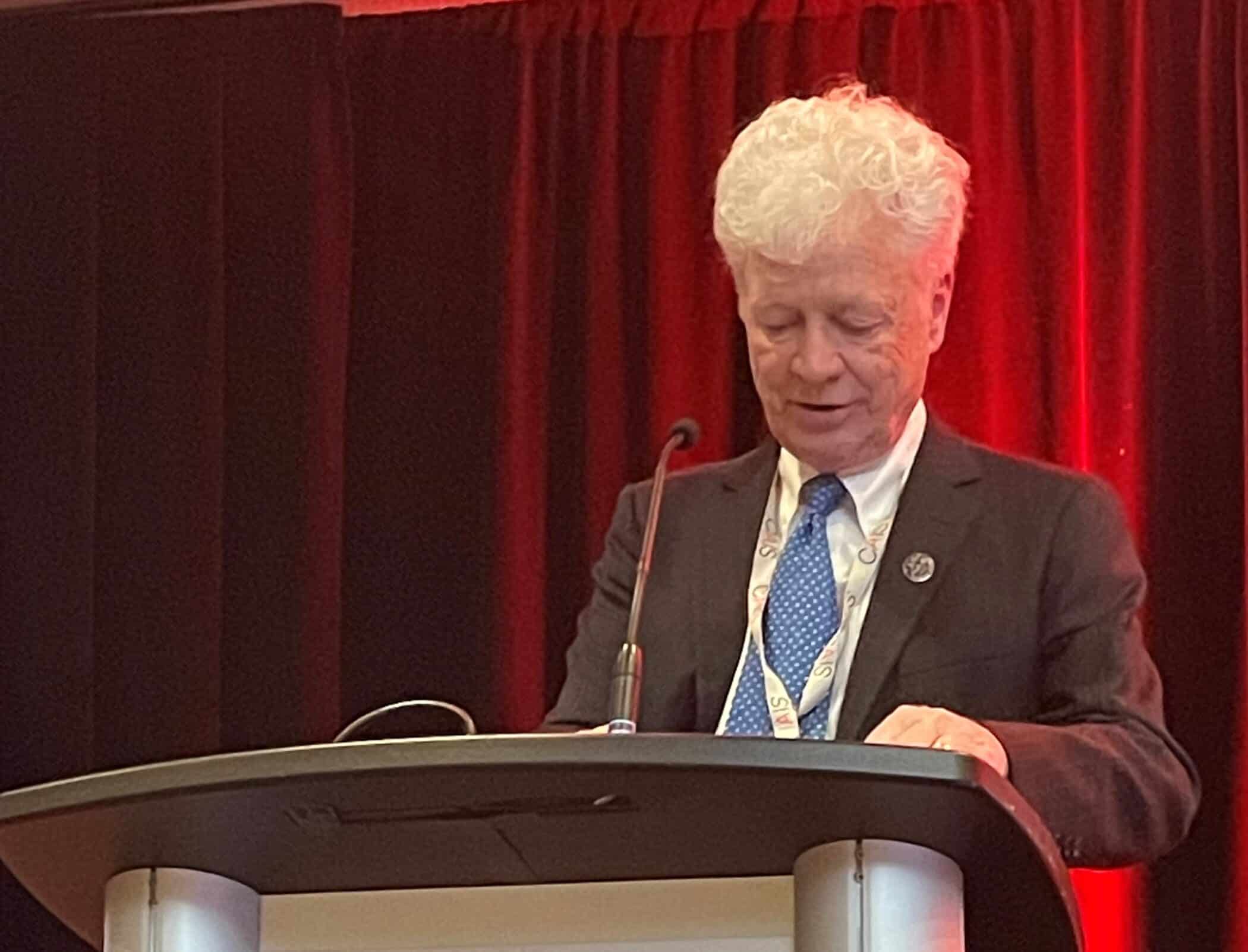An older man with white hair stands behind a lectern, reading from papers during a conference. Red curtains and CAIS branding form the backdrop.