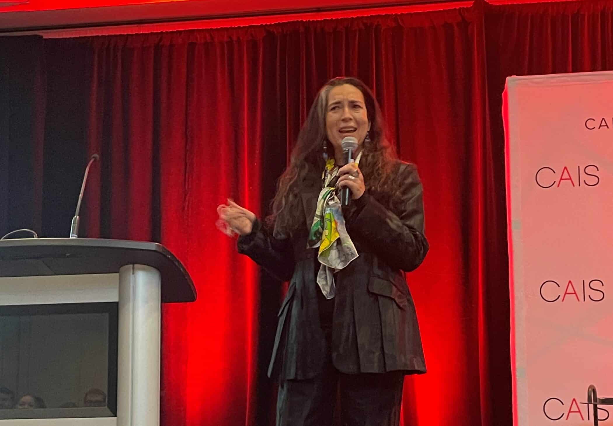 A woman speaks on stage holding a microphone in front of red curtains and a CAIS banner. She is wearing a dark suit with a colorful scarf and gesturing with her hand beside a lectern.