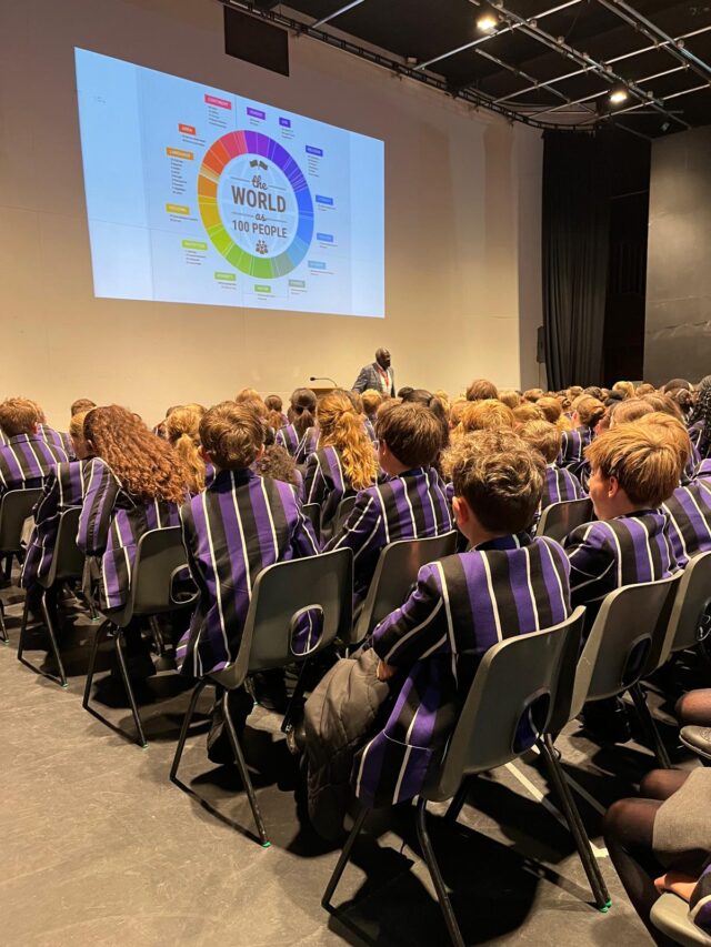 A large group of students wearing matching purple blazers with white stripes sit in rows in a school auditorium, facing a stage. A speaker stands at the front near a podium, addressing them. On the wall behind the speaker, a colorful circular infographic titled “The World as 100 People” is projected, showing various global demographic categories.