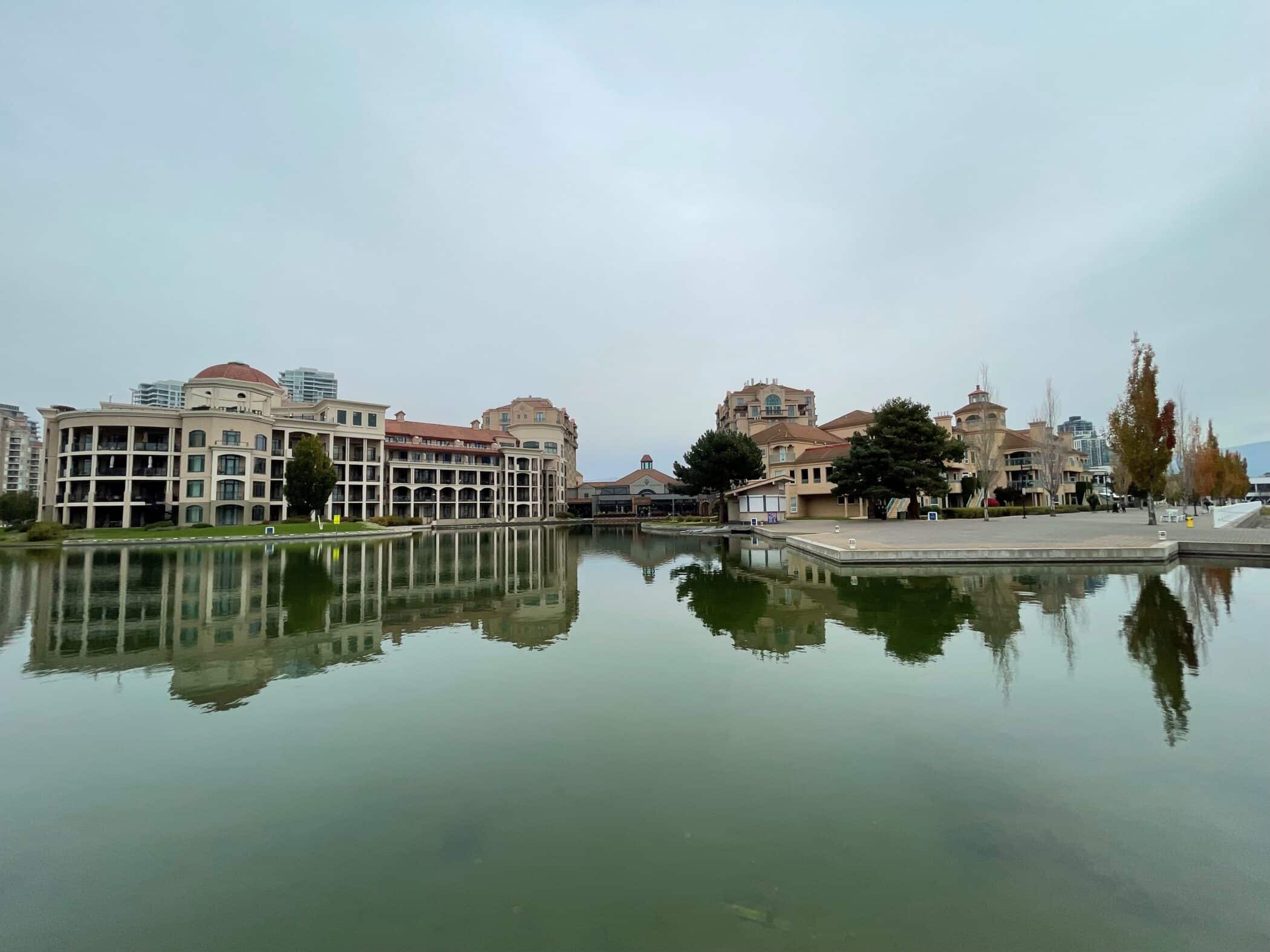 Waterfront view of the Kelowna Grand Okanagan Resort, its terracotta-roofed buildings reflected clearly in the still lagoon below.
