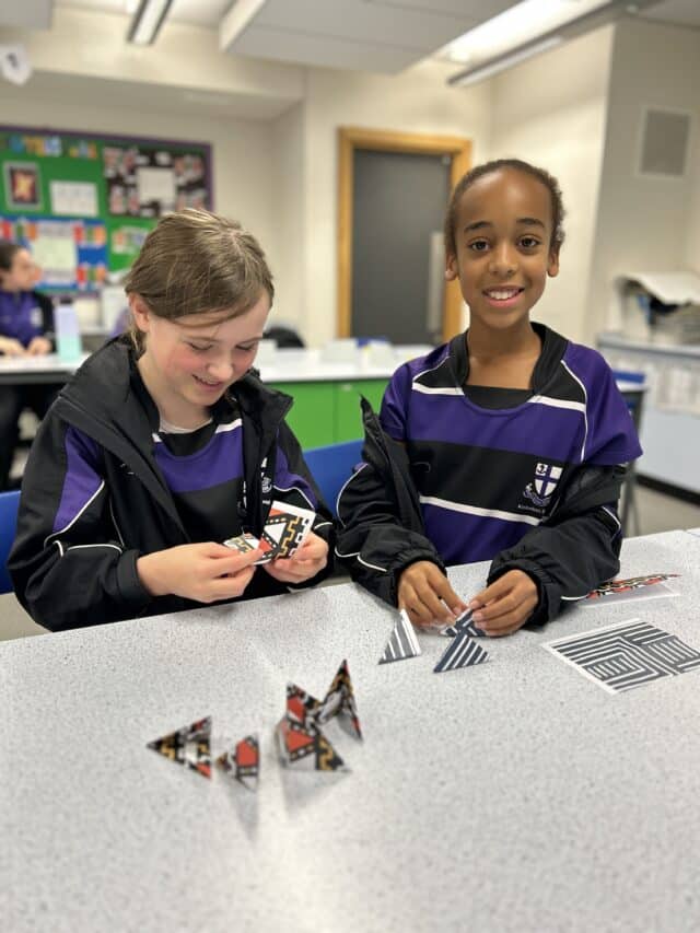 Two students in purple and black school uniforms sit at a classroom table working on a puzzle made of triangular patterned pieces. One student is smiling at the camera while the other looks down, focused on arranging the shapes. A colorful classroom display board is visible in the background.