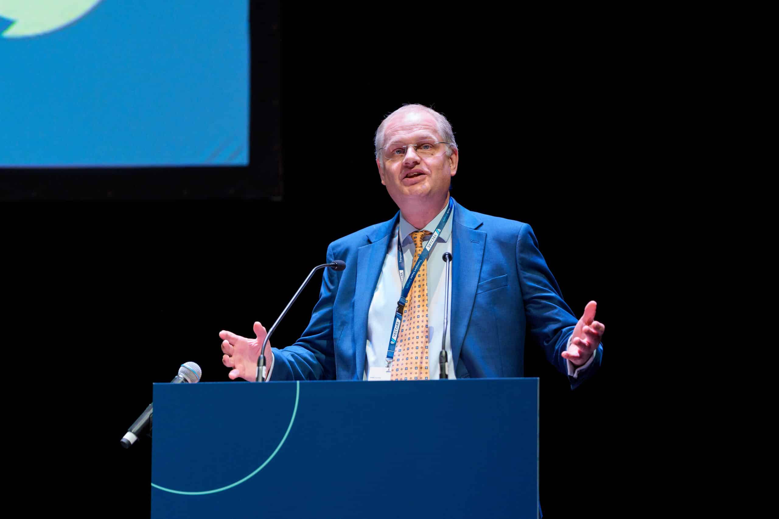 Philip Britton in a blue suit and patterned yellow tie speaks at a podium with microphones, gesturing with open hands, during the HMC Autumn Conference 2025 Opening Address