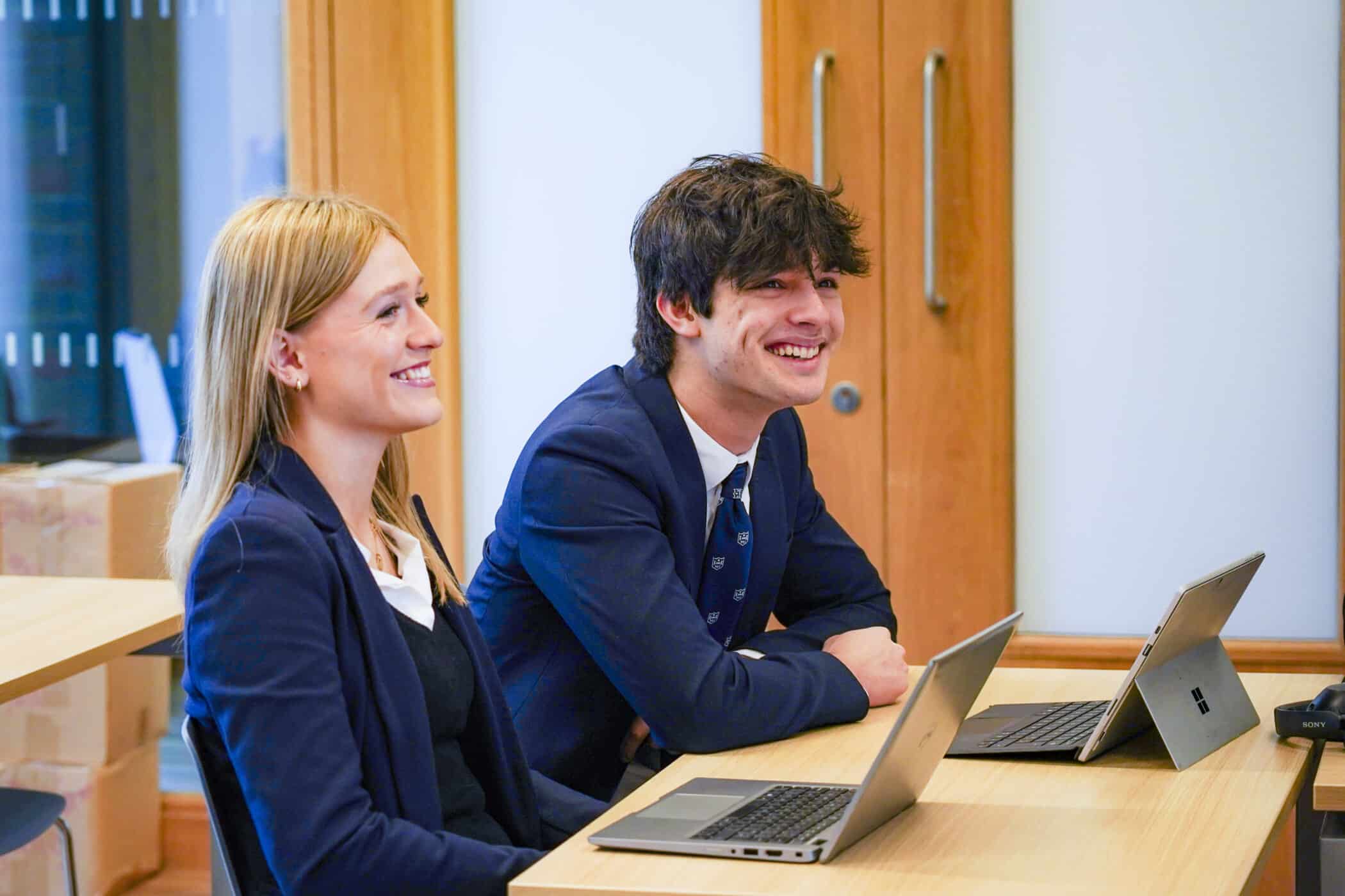 Two sixth-form students sit side by side at a desk in a modern classroom, smiling as they look towards the front. Both are dressed smartly in navy blazers, and each has a laptop open in front of them. The setting is bright and professional, with wooden panels and glass features in the background.