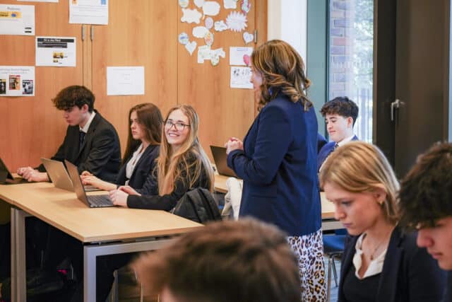 A classroom scene with sixth-form students using laptops at their desks. A teacher, dressed in a blazer, stands speaking to them while one student smiles at the camera. The room has wooden walls decorated with posters and paper cutouts, creating a warm and engaging learning environment.