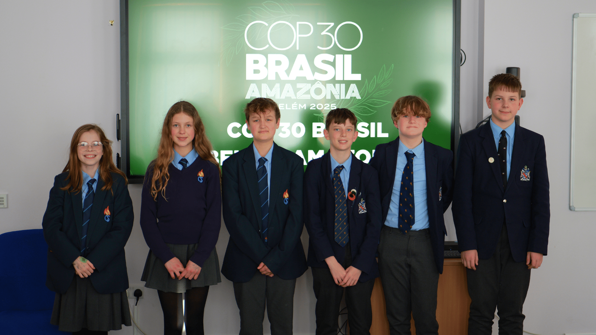Six schoolchildren in uniform stand in a row smiling in front of a large screen displaying the text “COP30 Brasil Amazônia Belém 2025.” They are dressed in blazers, shirts, ties, and skirts or trousers.