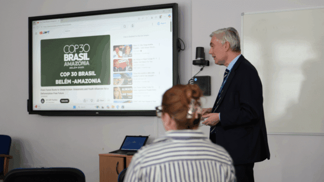 Melvyn Roffe, in a suit stands at the front of a classroom giving a presentation. A large screen behind him displays a YouTube page with a video titled “COP 30 Brasil Amazônia Belém 2025.” A person with their back to the camera listens, and a laptop sits on a small table beneath the screen. A whiteboard is visible on the wall to the right.