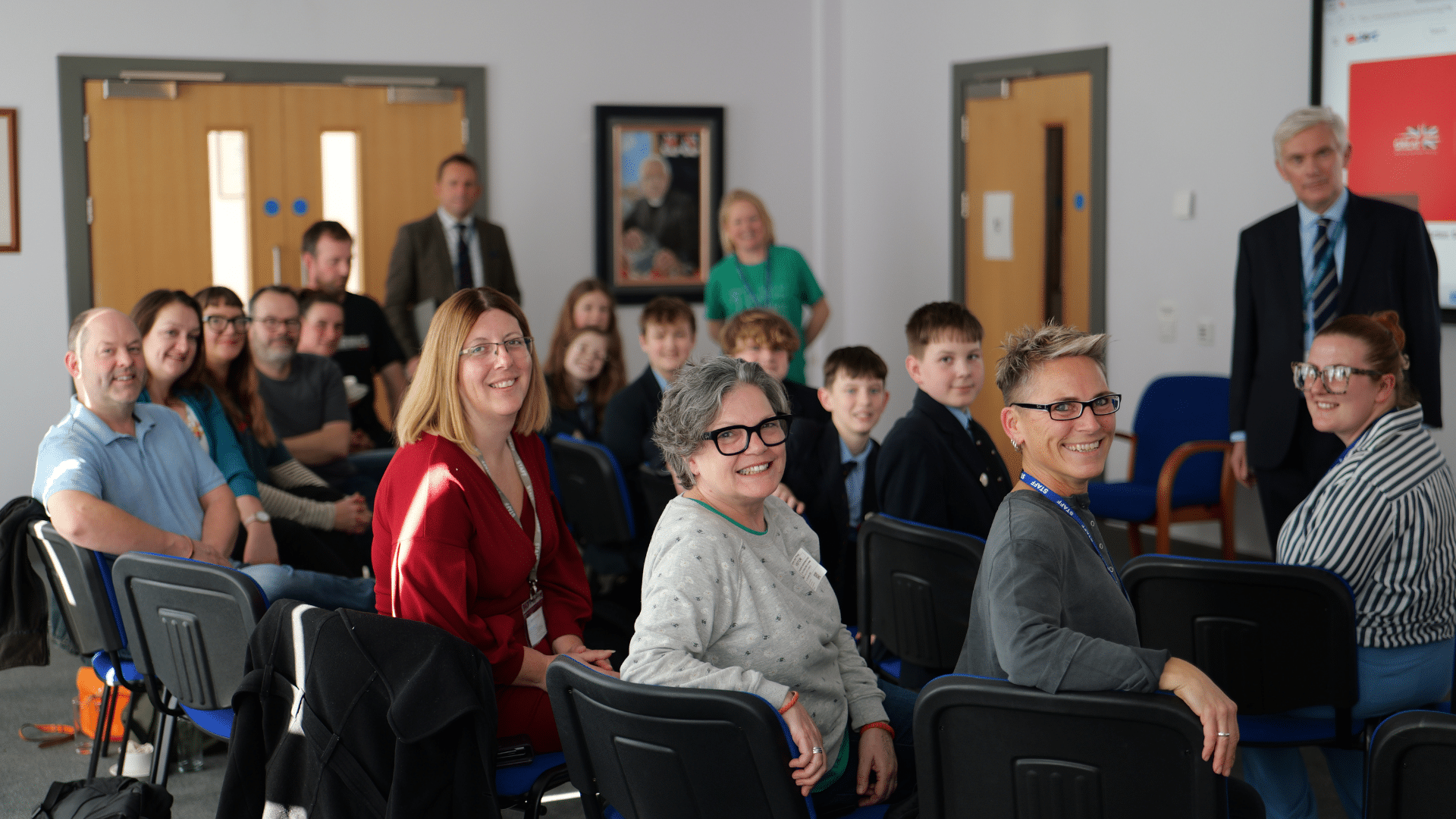 A group of adults and schoolchildren seated in rows of chairs in a classroom turn to smile at the camera. A man in a suit and a woman stand at the back of the room. The atmosphere appears friendly and informal.