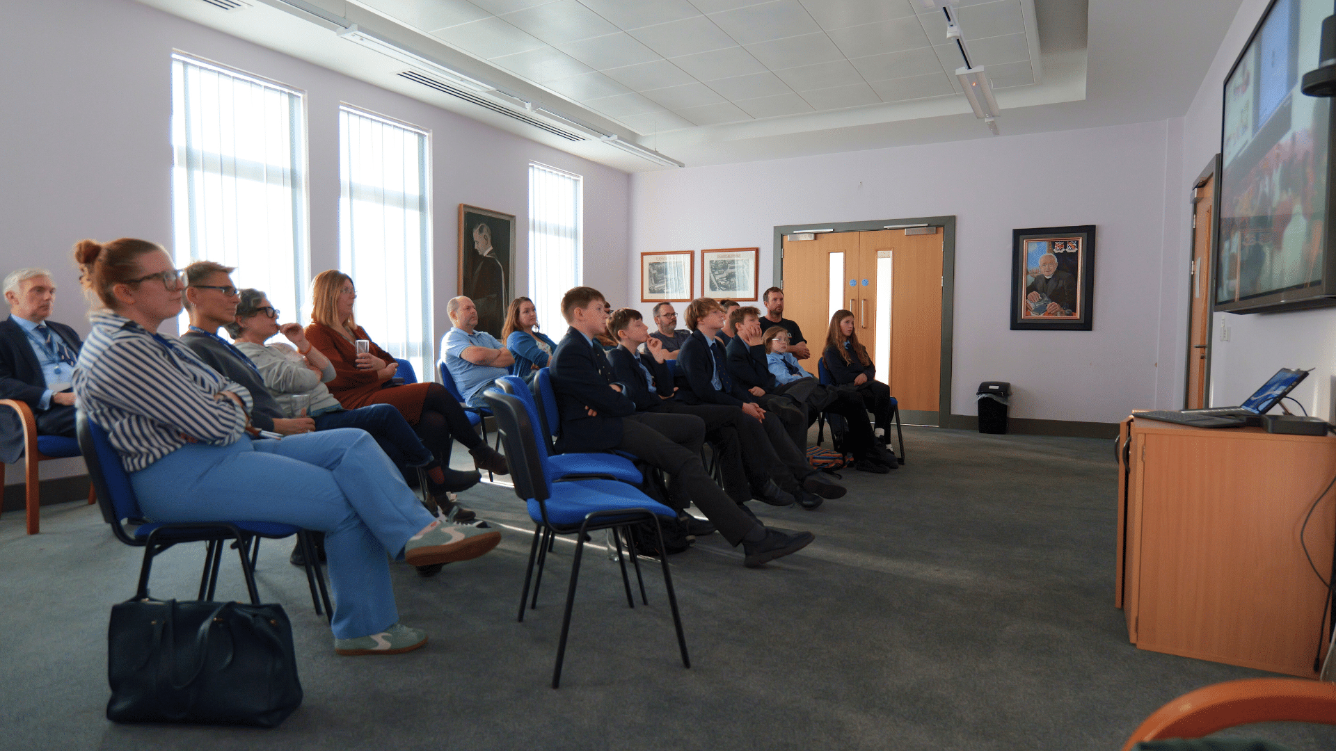 A mixed group of adults and schoolchildren sit in rows facing a large screen on the right side of the room, watching a presentation. Several framed artworks hang on the walls, and daylight streams through tall windows.