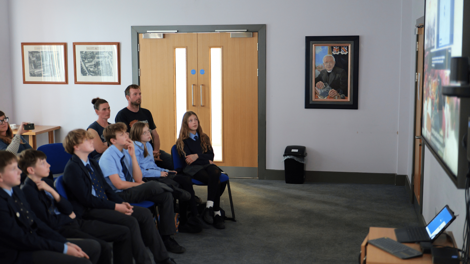 A group of schoolchildren sit in chairs watching a presentation on a large screen just out of frame to the right. Two adults sit behind them. A portrait and framed pictures hang on the wall near a set of double doors.