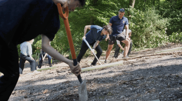 A group of people are outdoors on a wooded hillside, digging with shovels and spades. They appear to be working together on a planting or conservation activity under bright sunlight, surrounded by green trees.