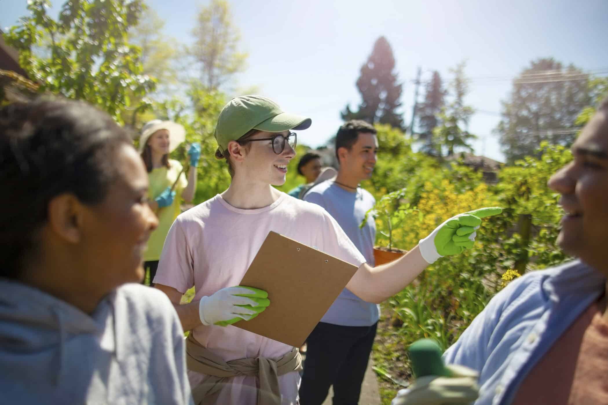 A group of volunteers works together in a sunny garden. At the center, a smiling young man wearing glasses, a green cap, and gardening gloves holds a clipboard and points toward another volunteer. People around him chat and carry plants and tools, surrounded by green foliage and bright sunlight.