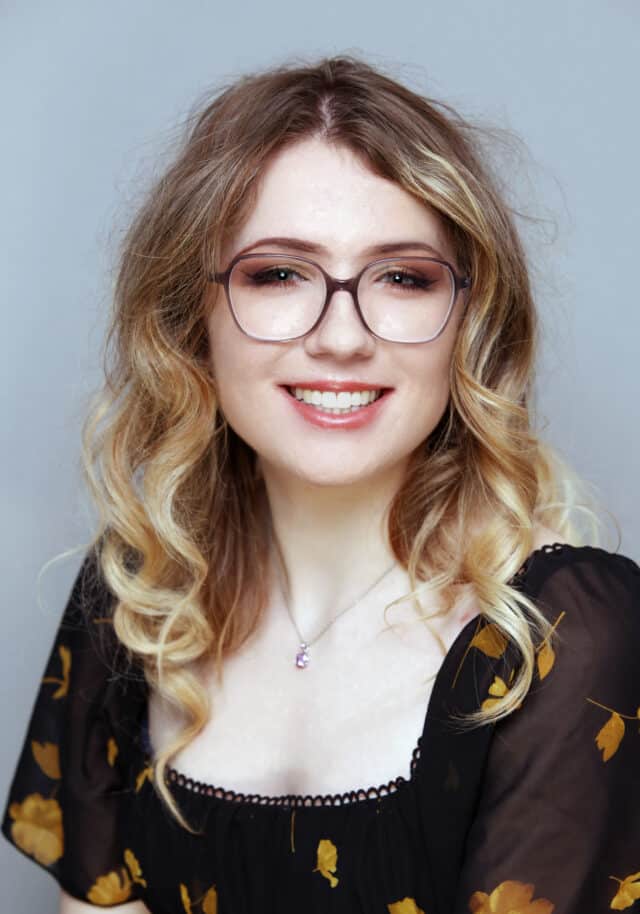 Smiling woman with wavy blonde hair wearing large round eyeglasses, a black dress with yellow floral print, and a small pendant necklace, posed against a plain light grey background.