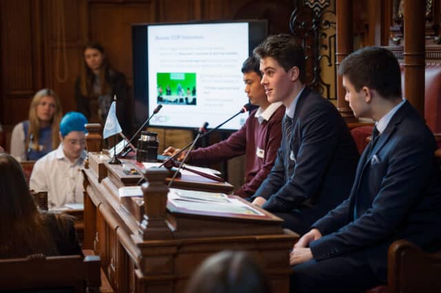 A group of three young men sit at a raised wooden bench during a formal conference. One in a suit speaks into a microphone, while another in a red jumper and one in a blue suit listen. Behind them, a screen displays a presentation slide titled “Recent COP Outcomes,” and several audience members watch attentively. The setting is a traditional wood-paneled hall with a serious, academic atmosphere.