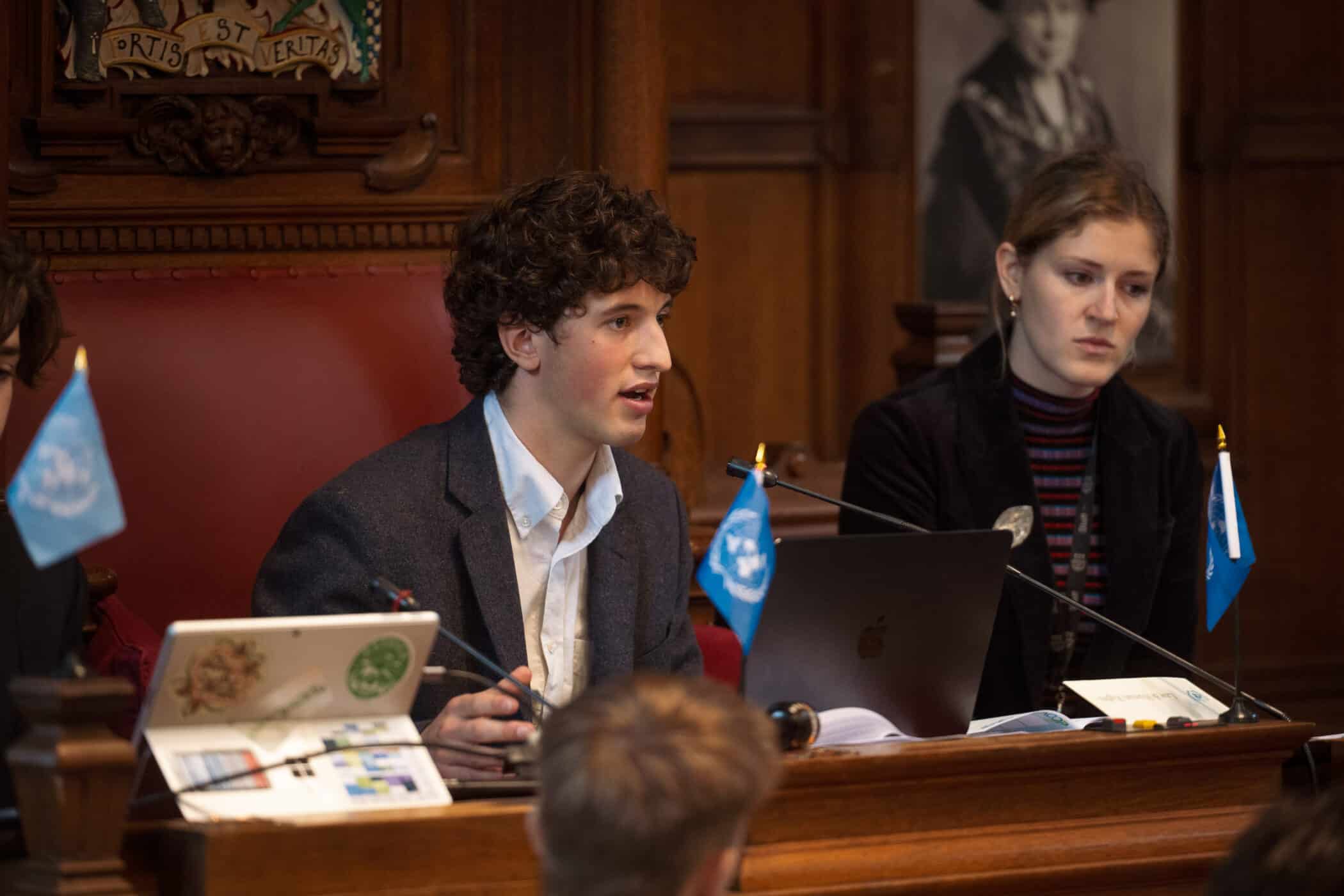 A young man with curly brown hair, dressed in a blazer and white shirt, speaks into a microphone while seated at a wooden desk in a grand, wood-panelled chamber. Next to him, a woman with blonde hair in a dark jacket listens attentively. Small United Nations flags are placed on the desk beside their laptops. The ornate woodwork behind them features a carved crest with the Latin words “Fortis Est Veritas.”