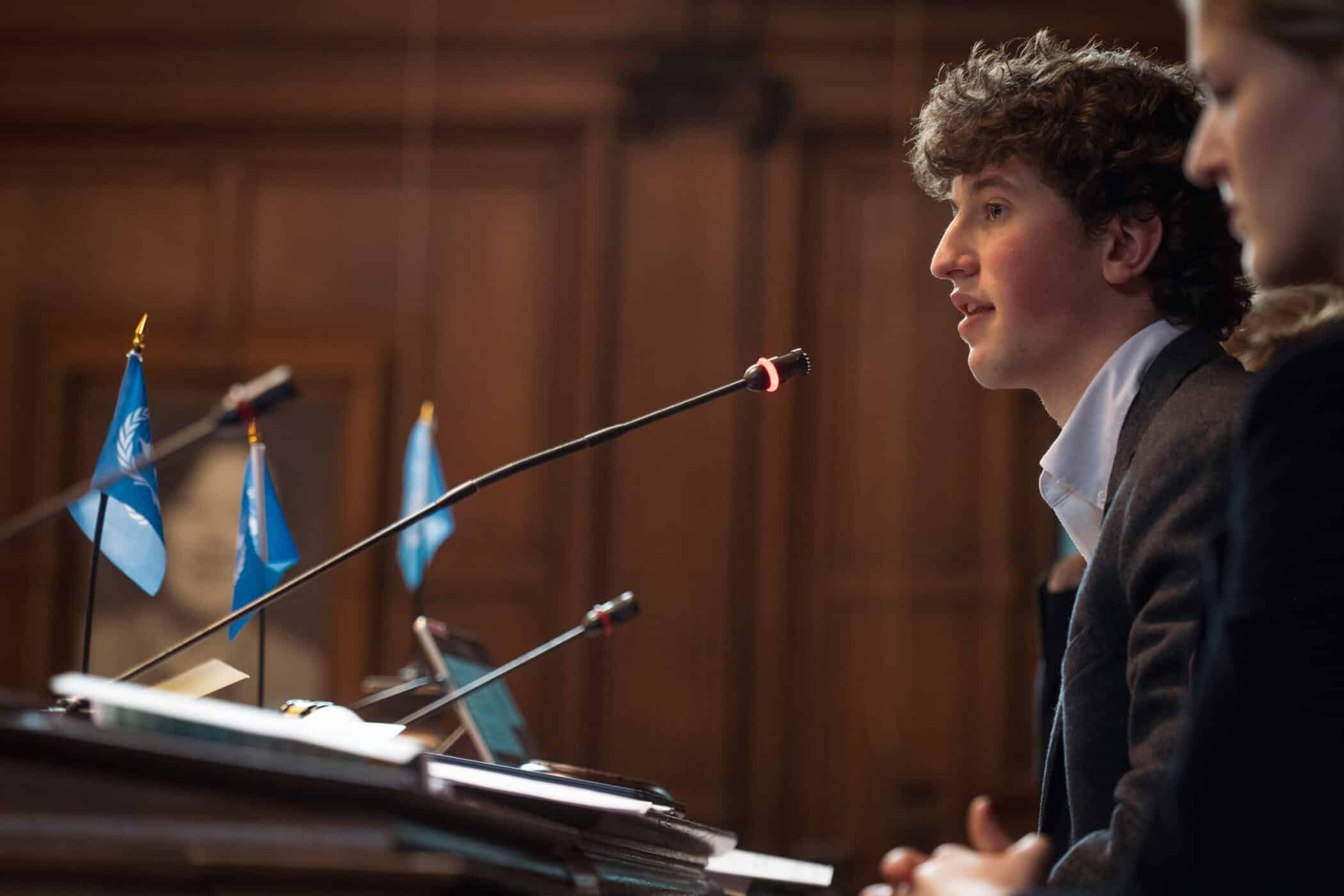 A side view of a young man with curly hair speaking passionately into a microphone at a formal desk. Blue United Nations flags line the desk in front of him. A woman sits beside him, slightly out of focus, while the background shows warm wooden paneling and a blurred portrait.