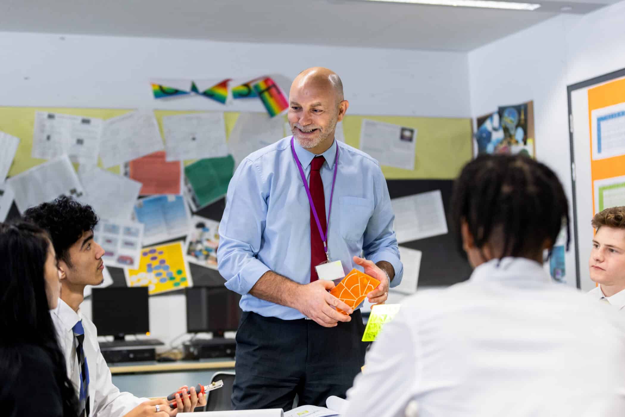 A smiling male teacher with a bald head and grey beard stands in front of a group of secondary school students in uniform. He holds colorful geometric teaching aids while the students listen attentively in a classroom decorated with posters and student work.