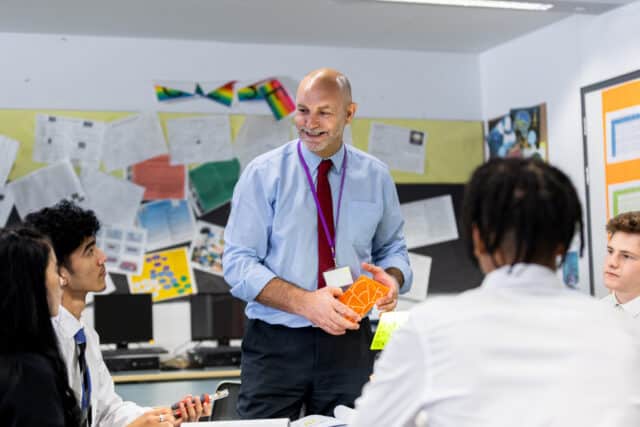A smiling male teacher with a bald head and grey beard stands in front of a group of secondary school students in uniform. He holds colorful geometric teaching aids while the students listen attentively in a classroom decorated with posters and student work.