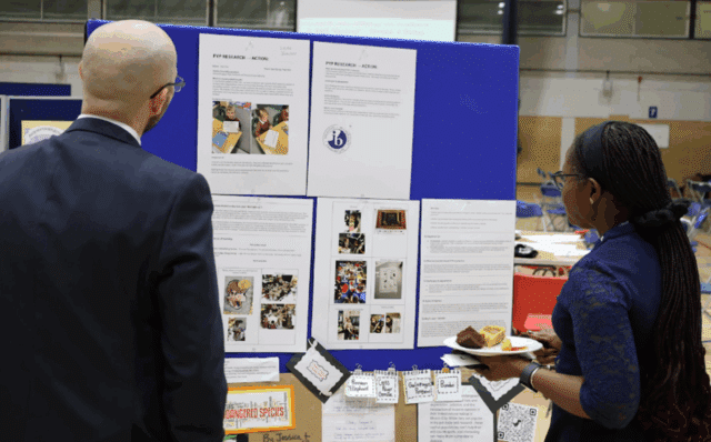 Two adults in professional attire view a blue display board featuring printed project materials, photos, and student work related to "PYP Research – Action." One person holds a plate with desserts while reading the display.