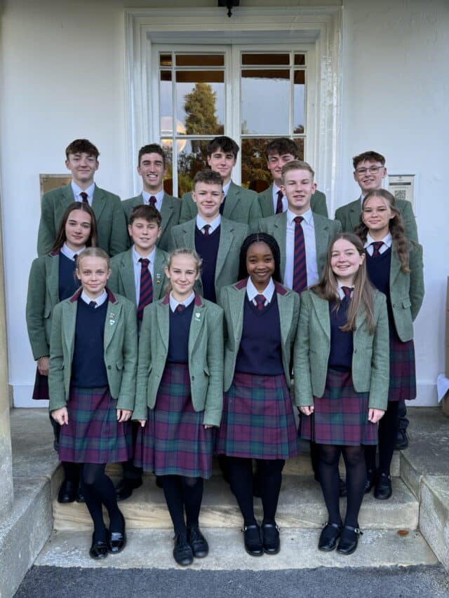 Group of 15 students in matching school uniforms, including green blazers and plaid skirts or trousers, standing on steps in front of a building with a white door and window.