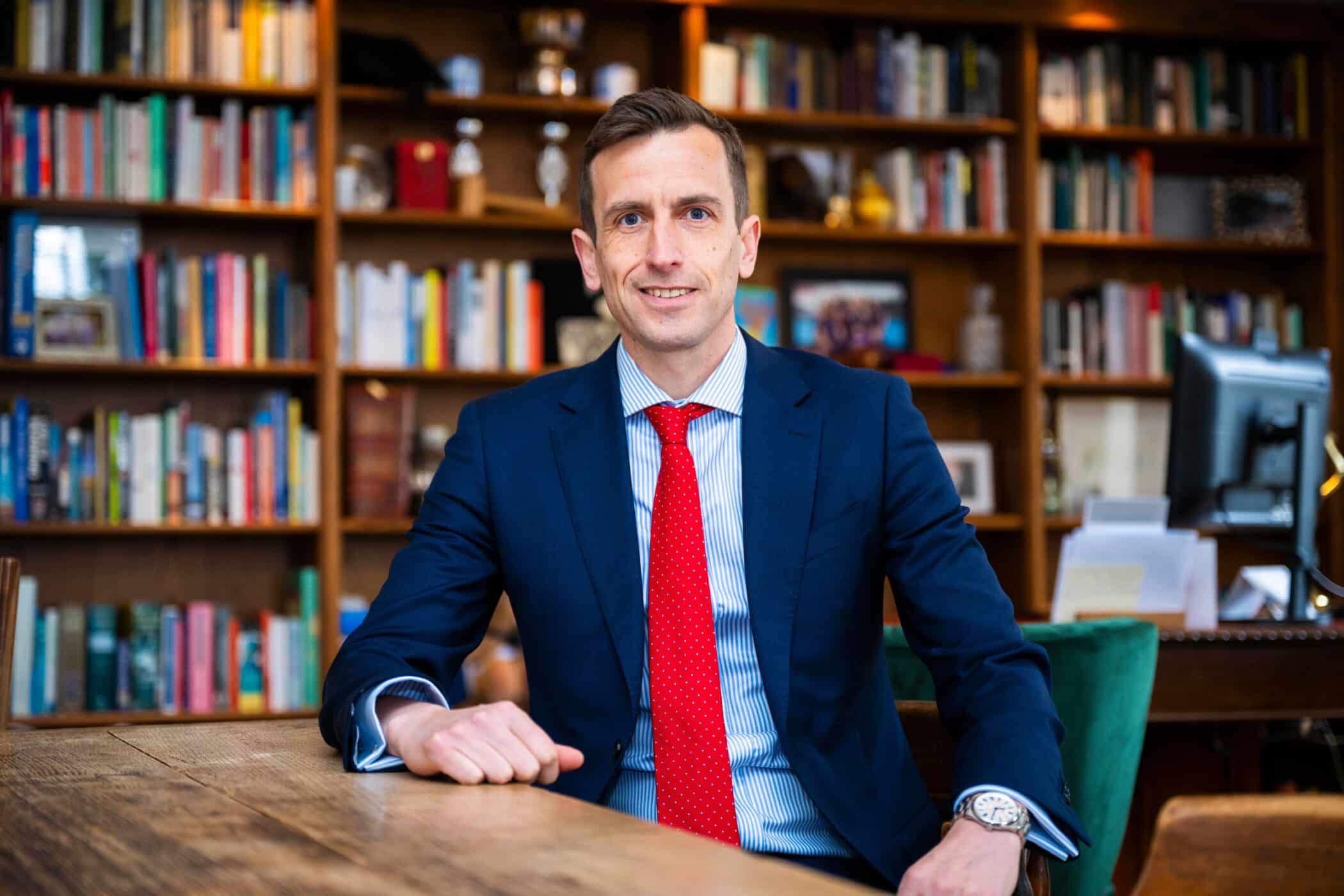 Man in a blue suit and red polka dot tie seated at a wooden table in a library or office, smiling. Shelves filled with books and framed photos are in the background.