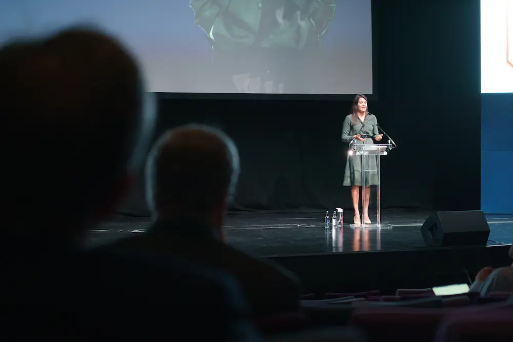 Woman standing at a lectern on a stage, speaking to an audience. A large screen is displayed behind her, with silhouetted attendees visible in the foreground of the auditorium.