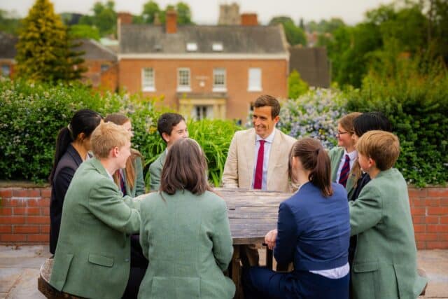 Man in a beige suit jacket and red tie sitting at a round outdoor table with a group of students in green blazers. They are engaged in conversation, with a school building and greenery in the background.
